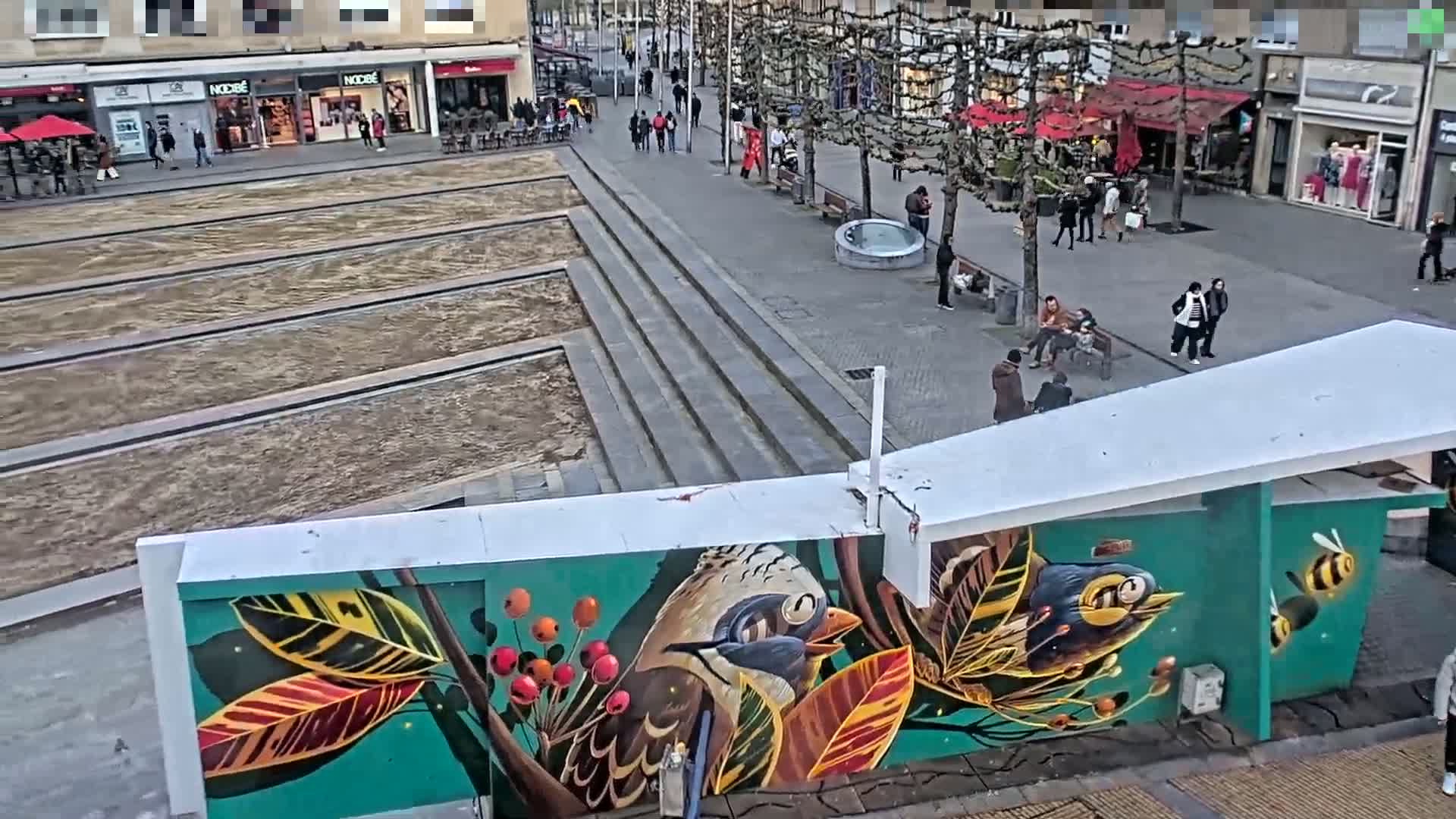 A mural depicting birds and a bee on a building wall is visible in a city square where several people are walking and sitting on benches under a partly cloudy sky.