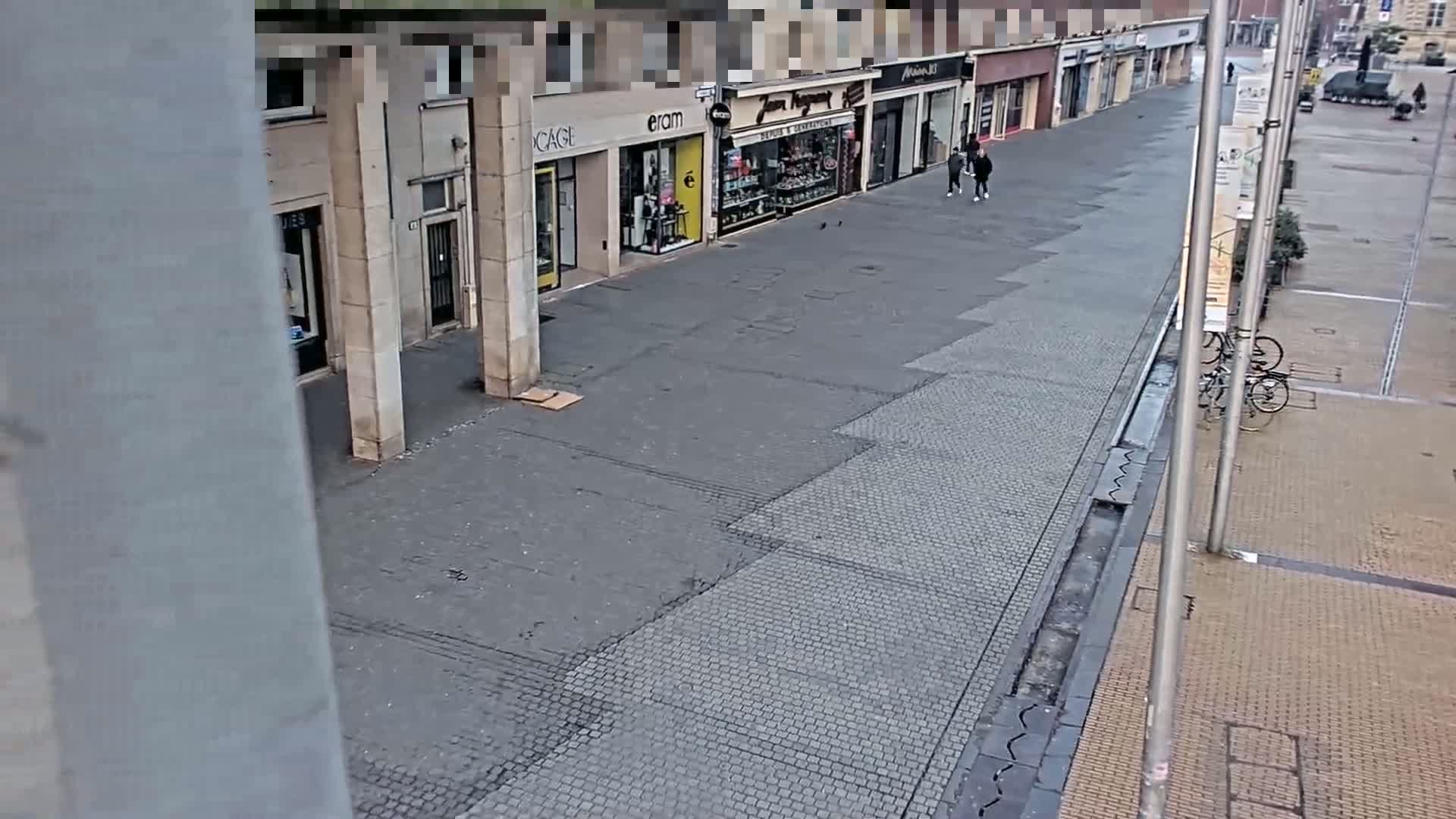A mostly empty paved pedestrian street lined with shops, with a few people walking and bicycles parked nearby, on an overcast day.