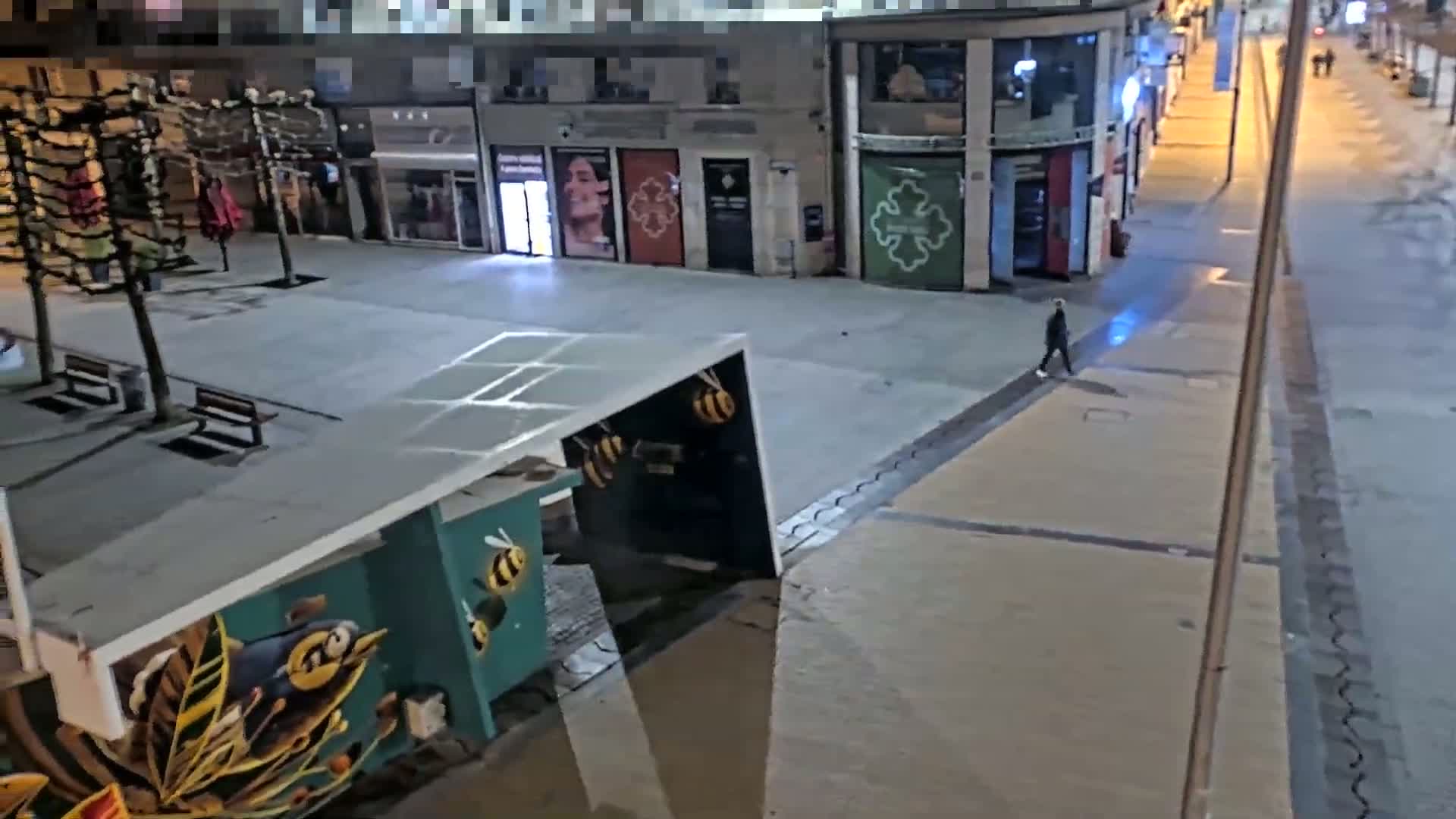 A lone person walks down a mostly empty, well-lit city street at night.
