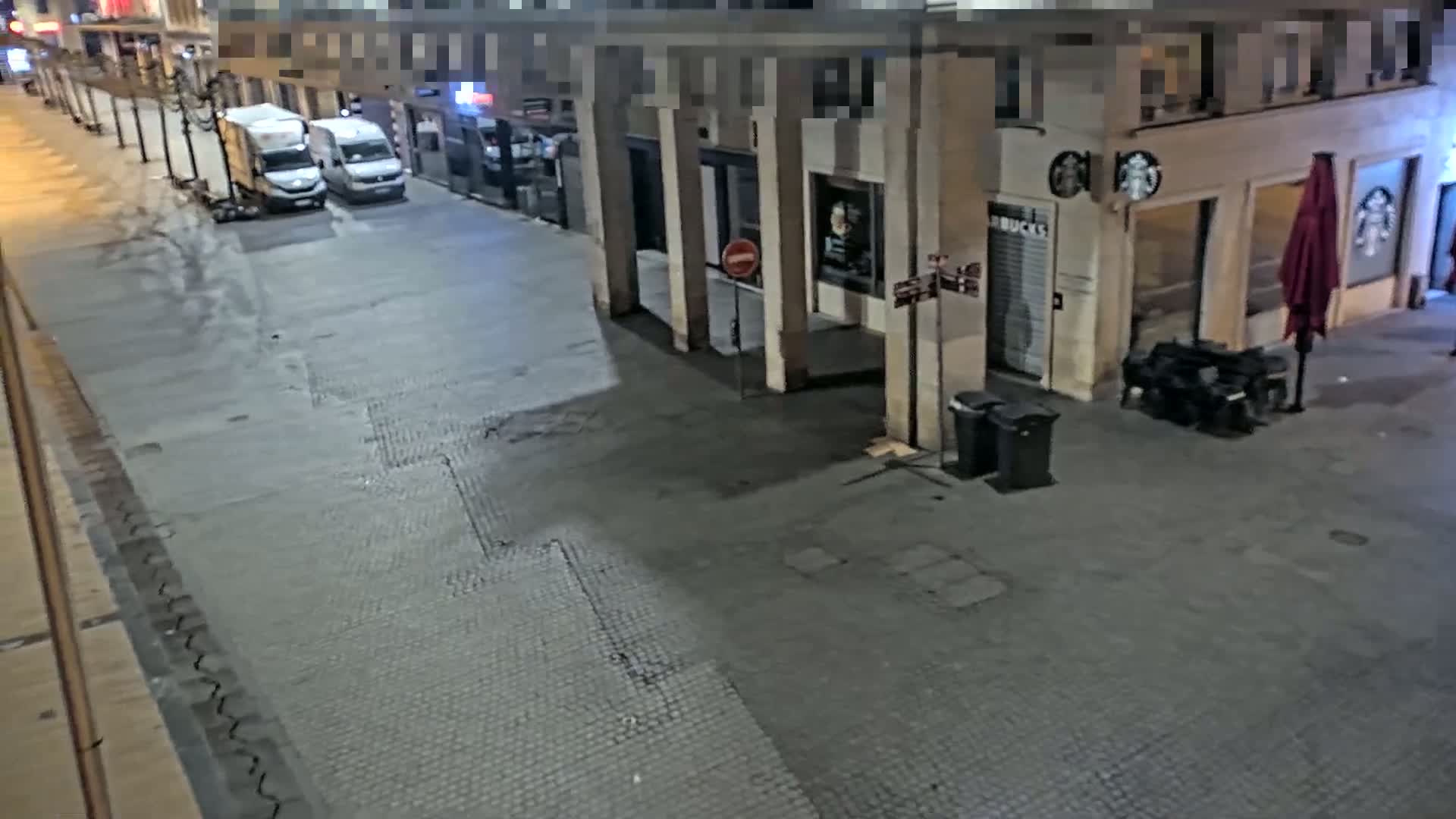 A nighttime, empty street scene shows two delivery vans parked near a Starbucks, with some trash cans and a closed red umbrella nearby.