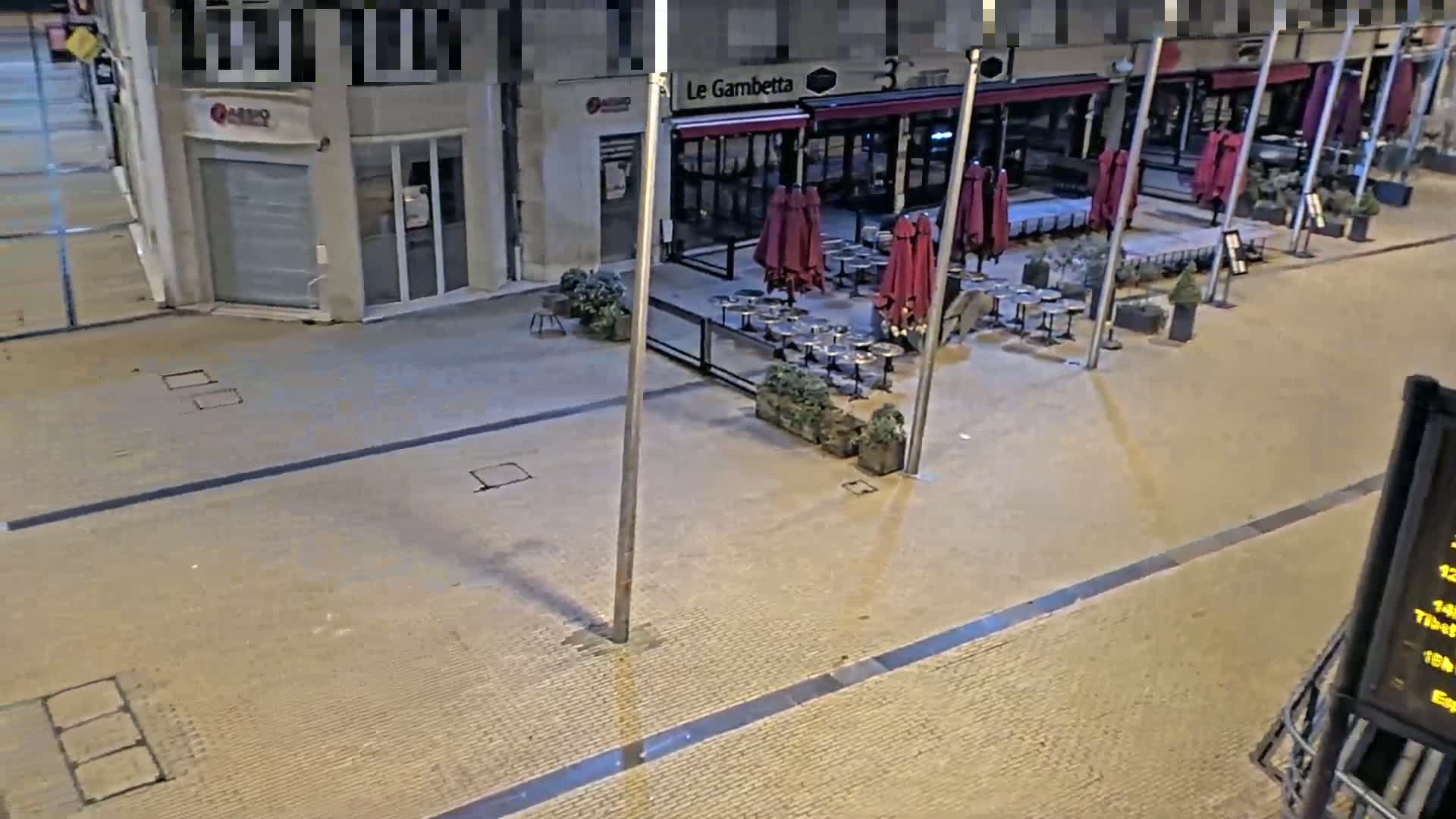 An empty outdoor cafe with maroon umbrellas and metal tables sits on a paved plaza at night.