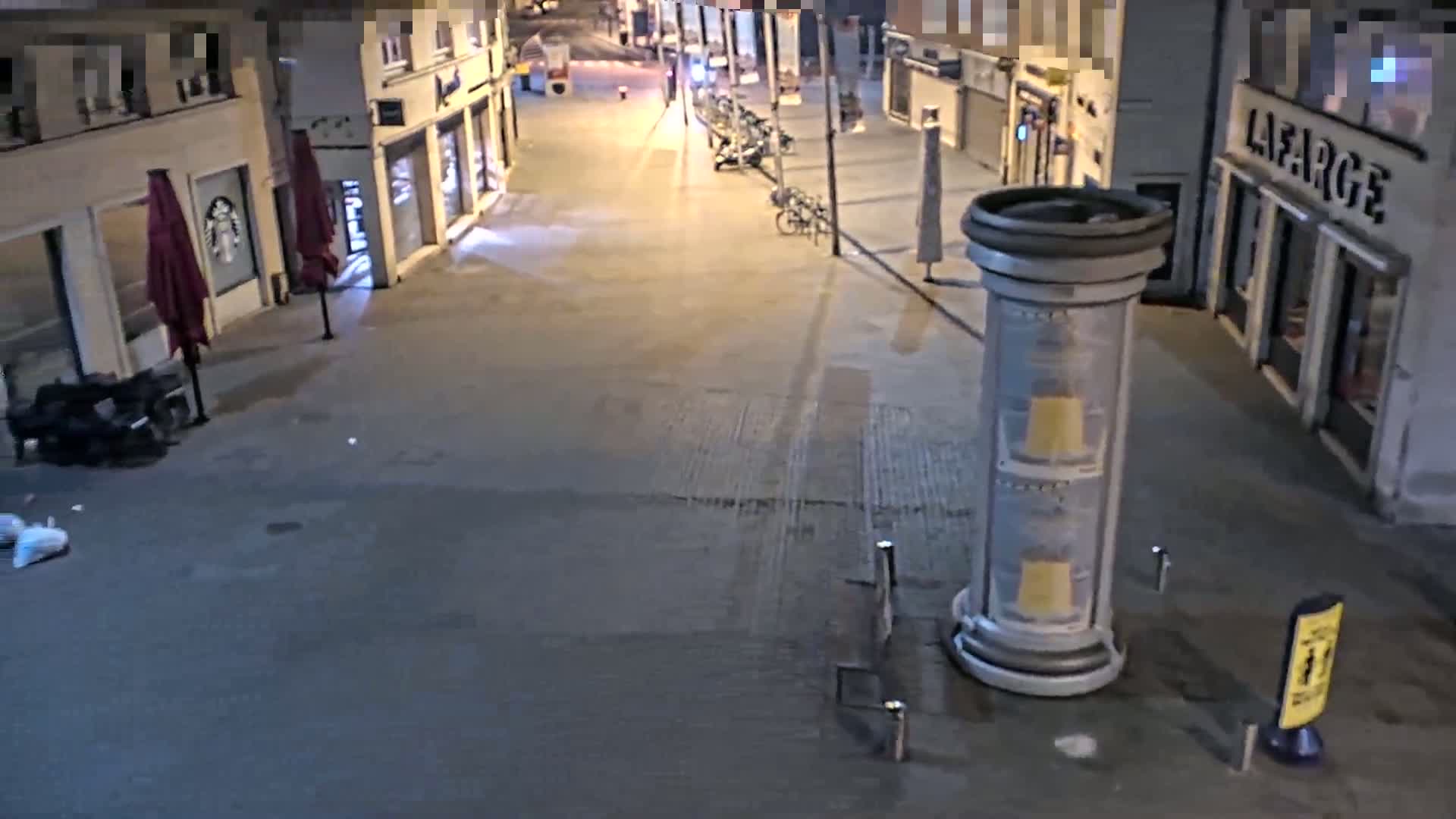 A nighttime street scene shows an empty city block with parked bicycles, closed shops, and a large advertising column.