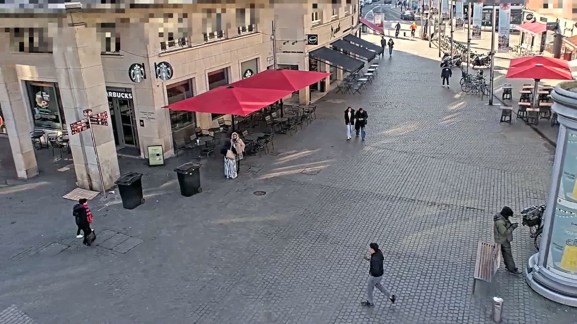 A cobblestone plaza with a Starbucks and several outdoor cafes under red umbrellas, and a few pedestrians are visible on a sunny day.