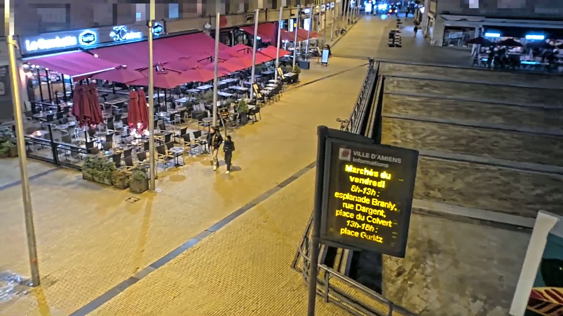 A wet, paved city street at night shows an outdoor cafe with red umbrellas and several occupied tables, and two pedestrians walking nearby.