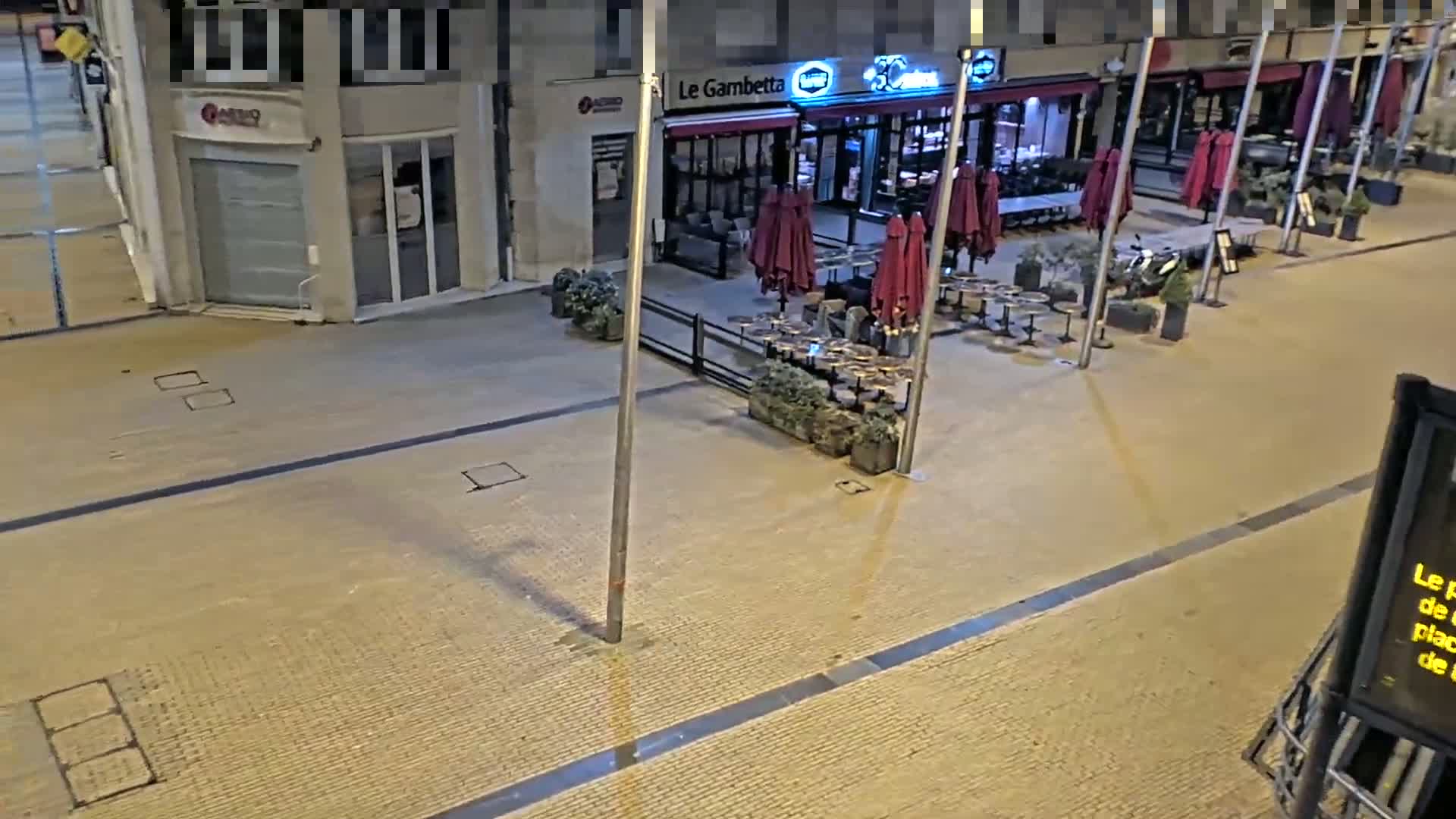 An empty, paved city street at night features a restaurant with closed maroon umbrellas and outdoor seating.