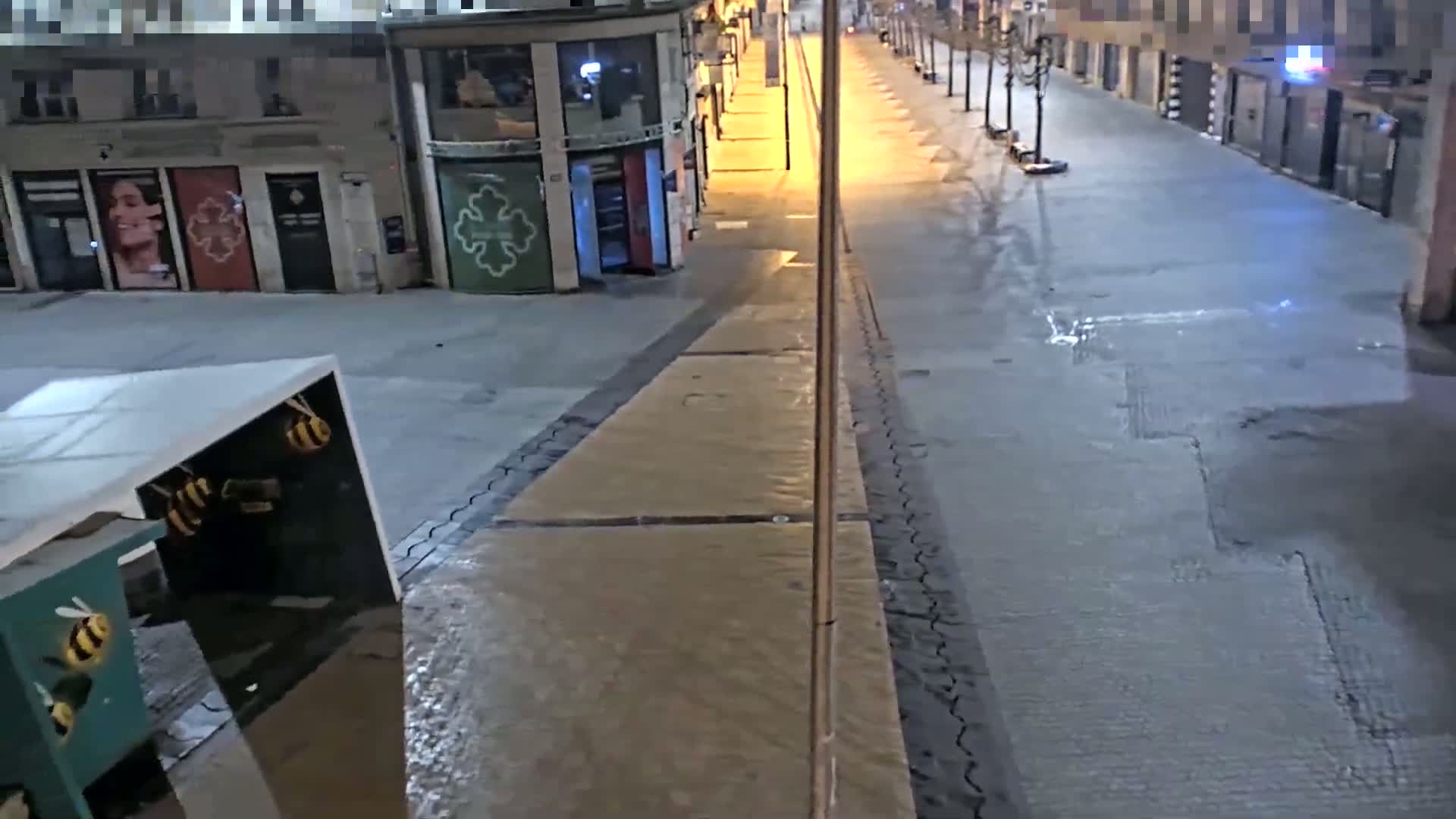 A mostly empty, wet street at night is lit by streetlights, with buildings lining both sides.