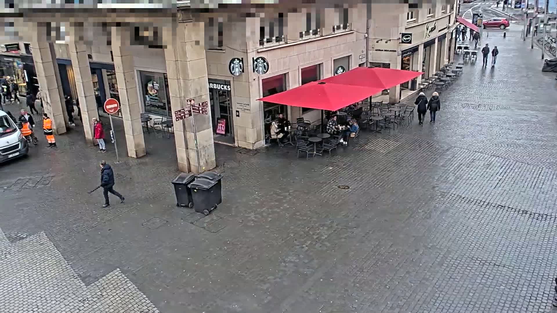 A wet cobblestone street scene shows people walking past a Starbucks with outdoor seating under a red umbrella.