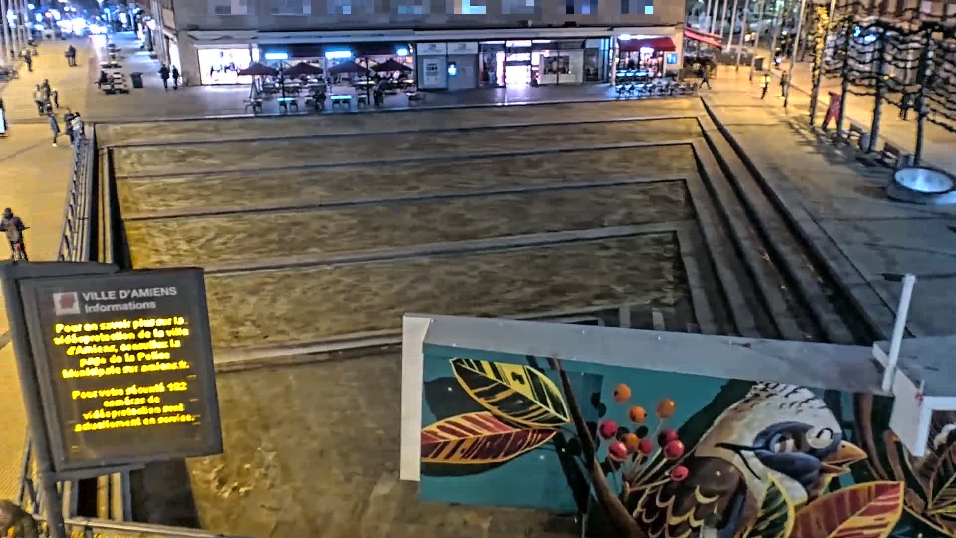 A nighttime view of a city square with a long, shallow, rectangular water feature, and a mural of a bird and leaves on a wall.