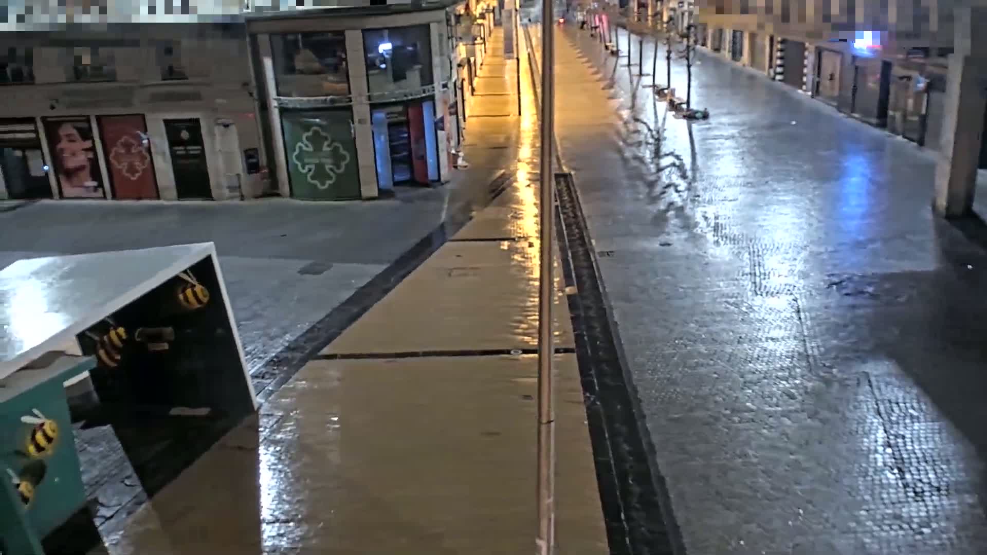 A wet, empty city street at night is lit by streetlights, with buildings lining both sides.