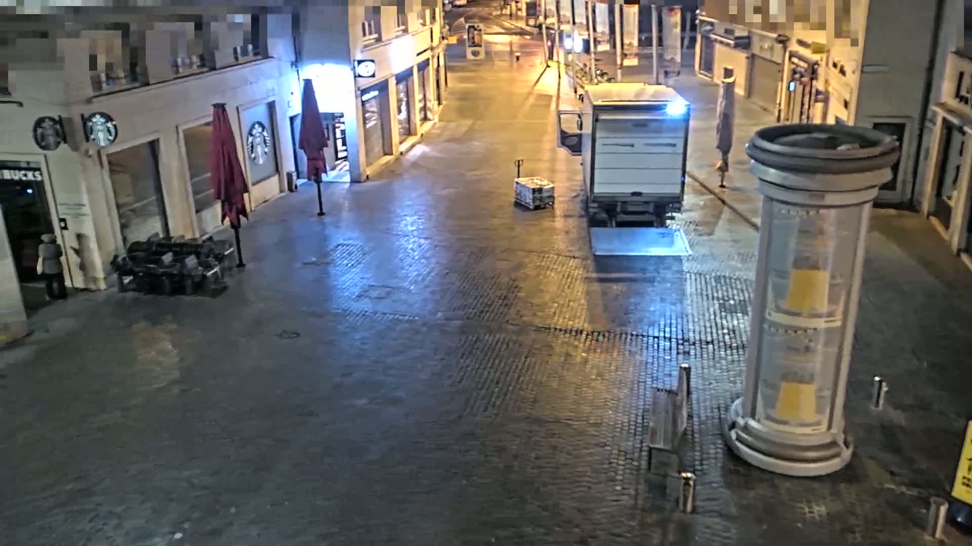 A mostly empty cobblestone street at night is lit by streetlights, with a white delivery truck parked near a Starbucks and a tall, cylindrical sign.
