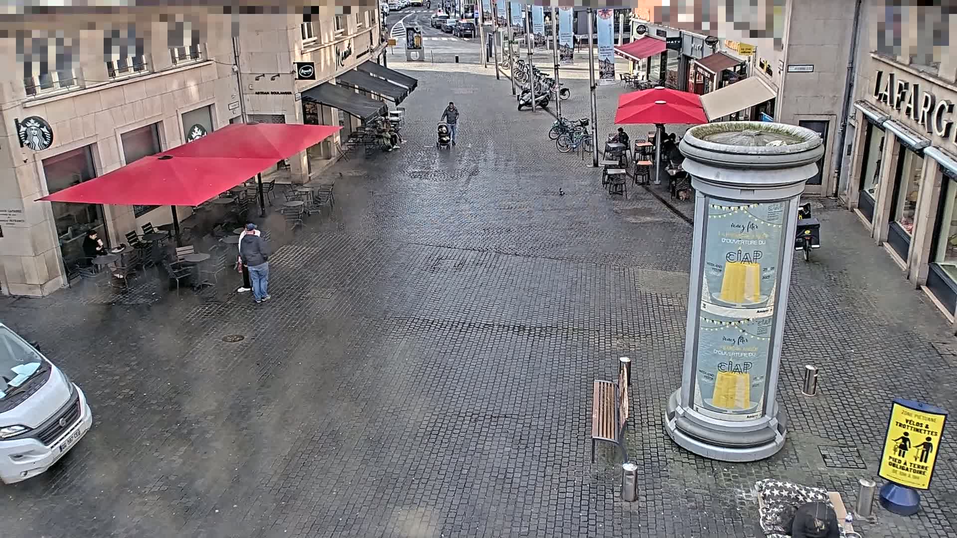 A cobblestone city square with several outdoor café seating areas under red umbrellas, a few pedestrians, and bicycles parked nearby, on a cloudy day.