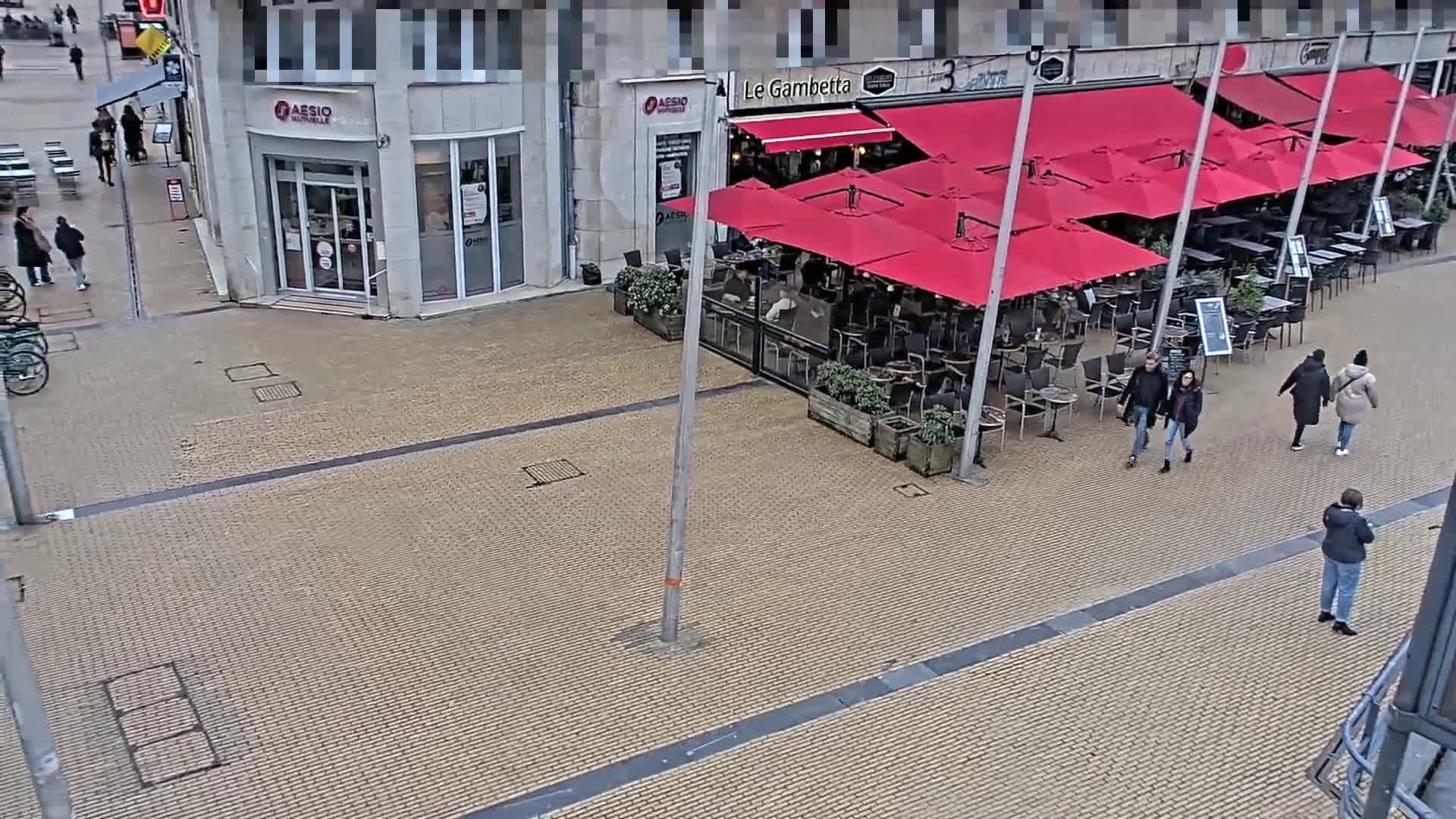 An outdoor paved area shows a restaurant with red umbrellas and several people walking around on an overcast day.