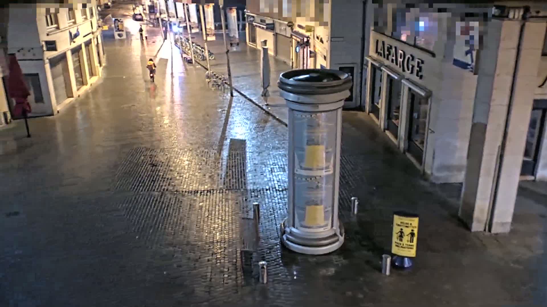 A wet, cobblestone street at night is seen with a person on a bicycle, parked bicycles along the side of the street, and a tall, cylindrical advertisement stand.