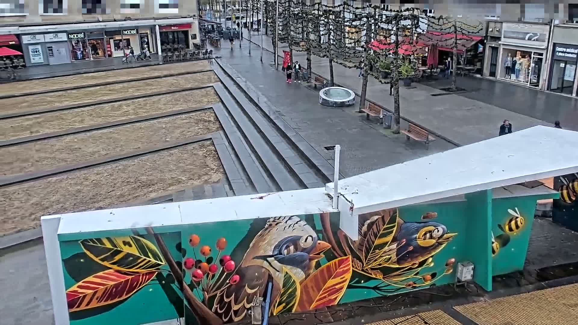 A mural depicting birds and bees on a teal background is visible beneath a white overhang in a city square on an overcast day.