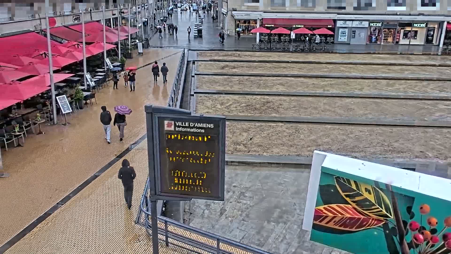 A wet city square with red umbrellas outside a cafe, pedestrians walking, and a large, empty planter area.
