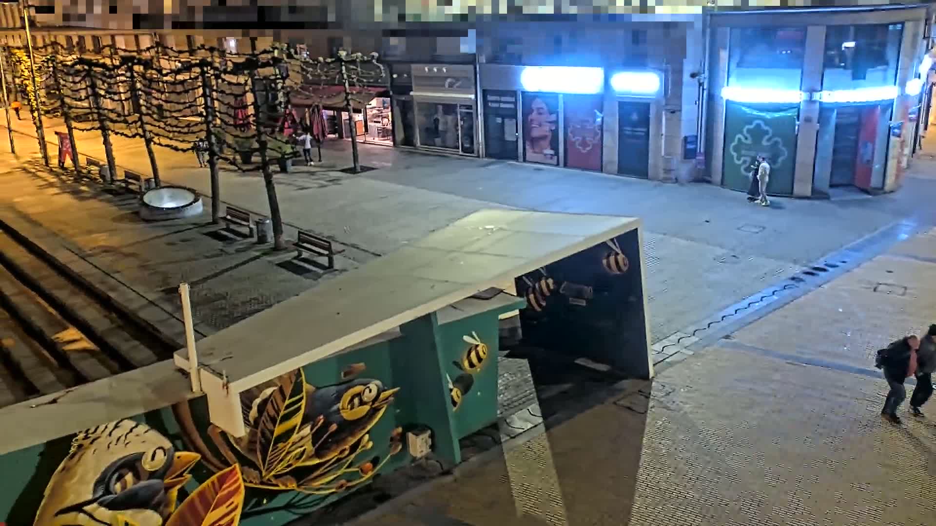 A nighttime city square scene shows a few people walking near shops and a bus shelter with bee-themed artwork, under the cover of darkness.