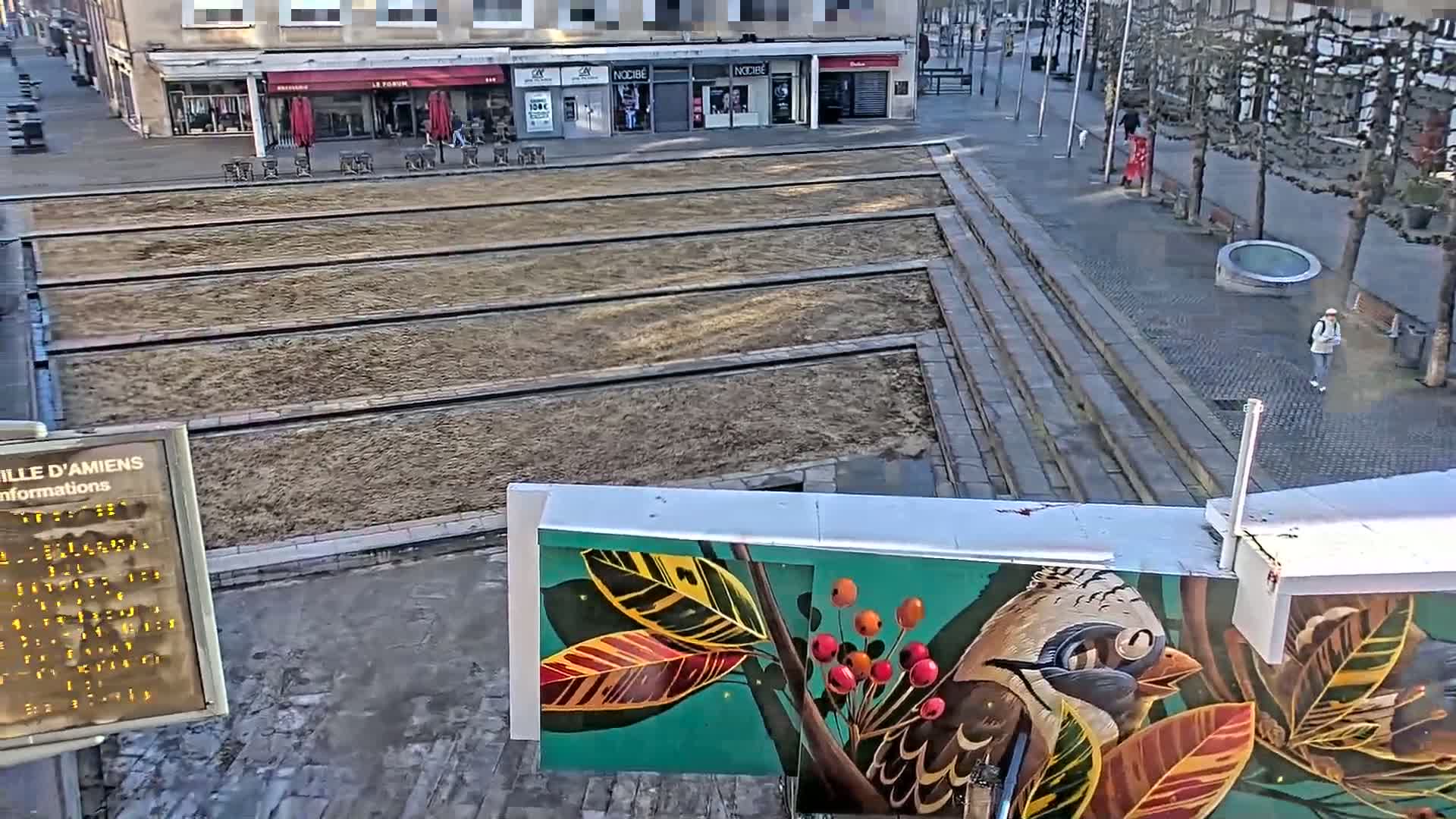 A mostly sunny view of a town square with a large, rectangular, barren area divided into sections by raised lines, and a mural depicting a bird and leaves on a building.