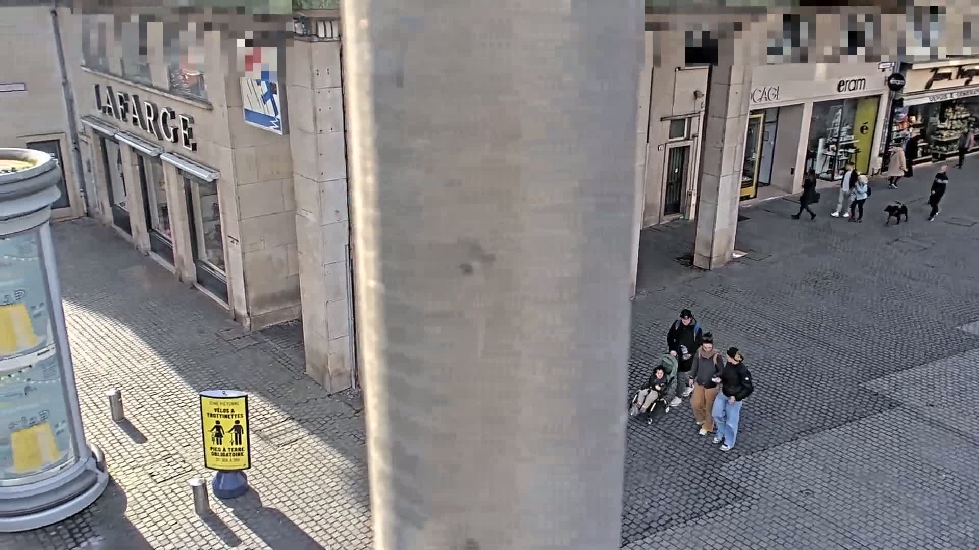 A high-angle view shows a paved city street with several pedestrians, some walking in groups, on a sunny day.