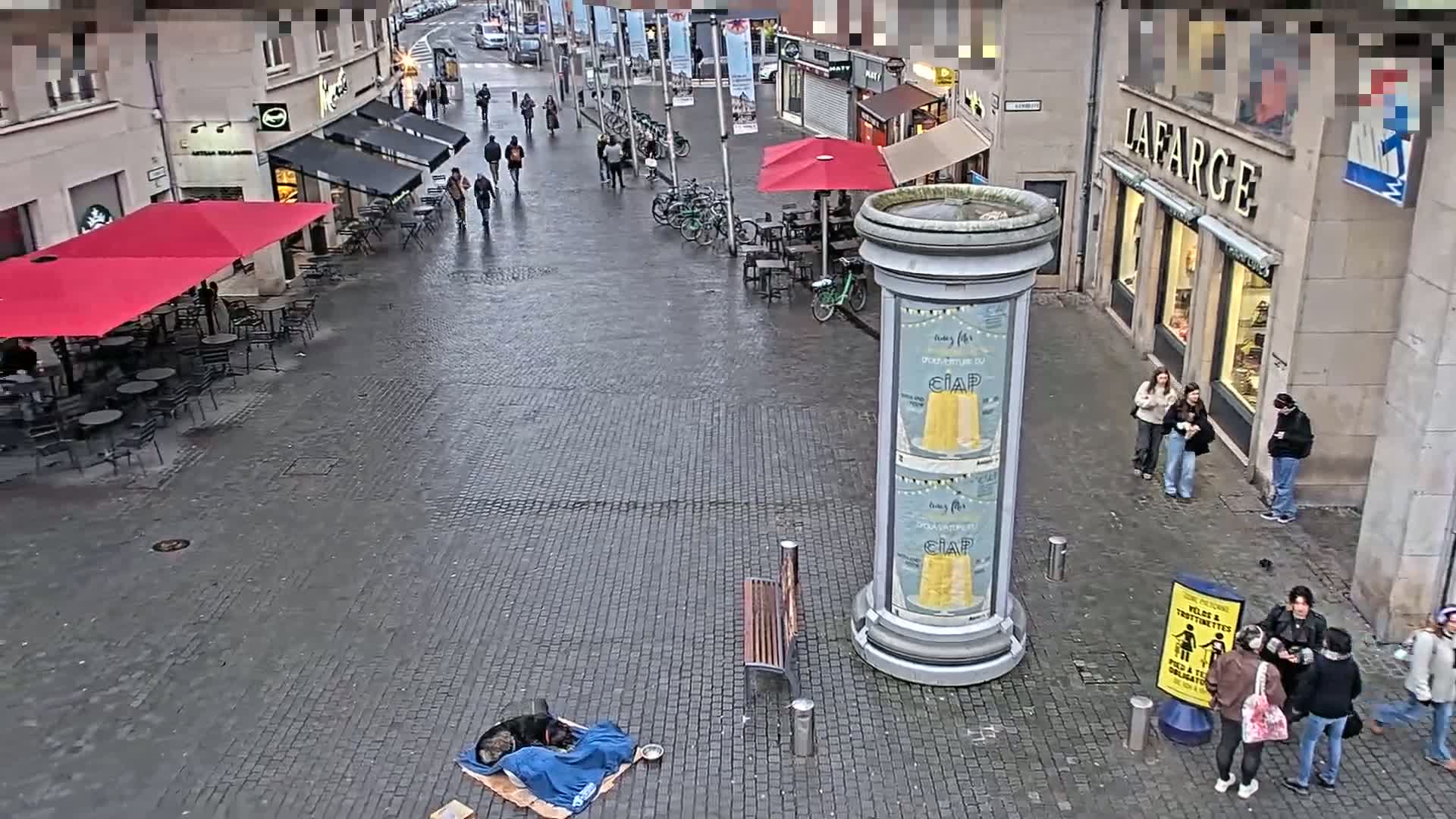 A wet cobblestone street scene shows several pedestrians, outdoor seating areas with red umbrellas, bicycles, and a person sleeping on the ground under a blanket.