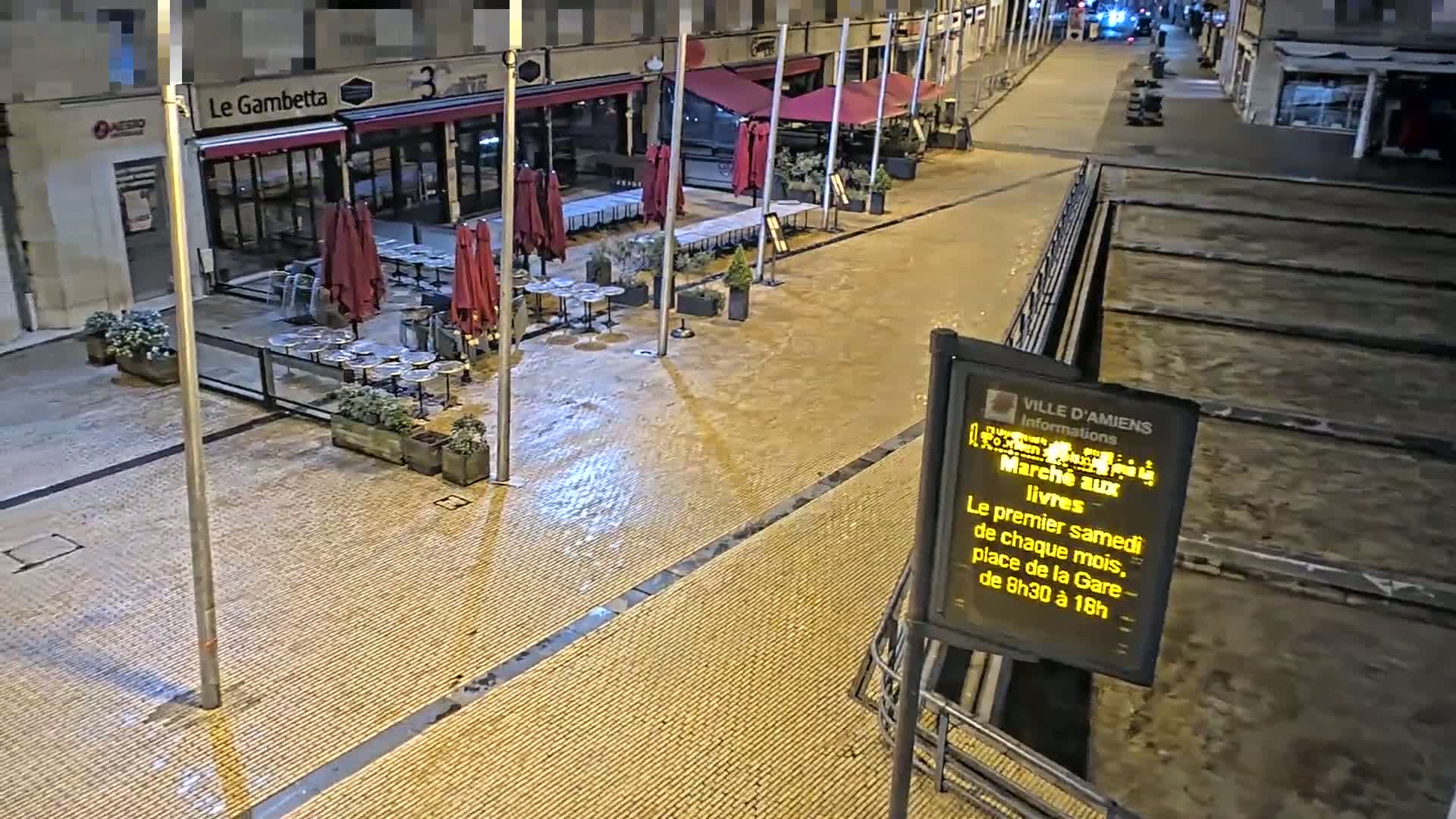 A wet, nighttime street scene shows several closed restaurants with outdoor seating areas, and a mostly empty plaza.