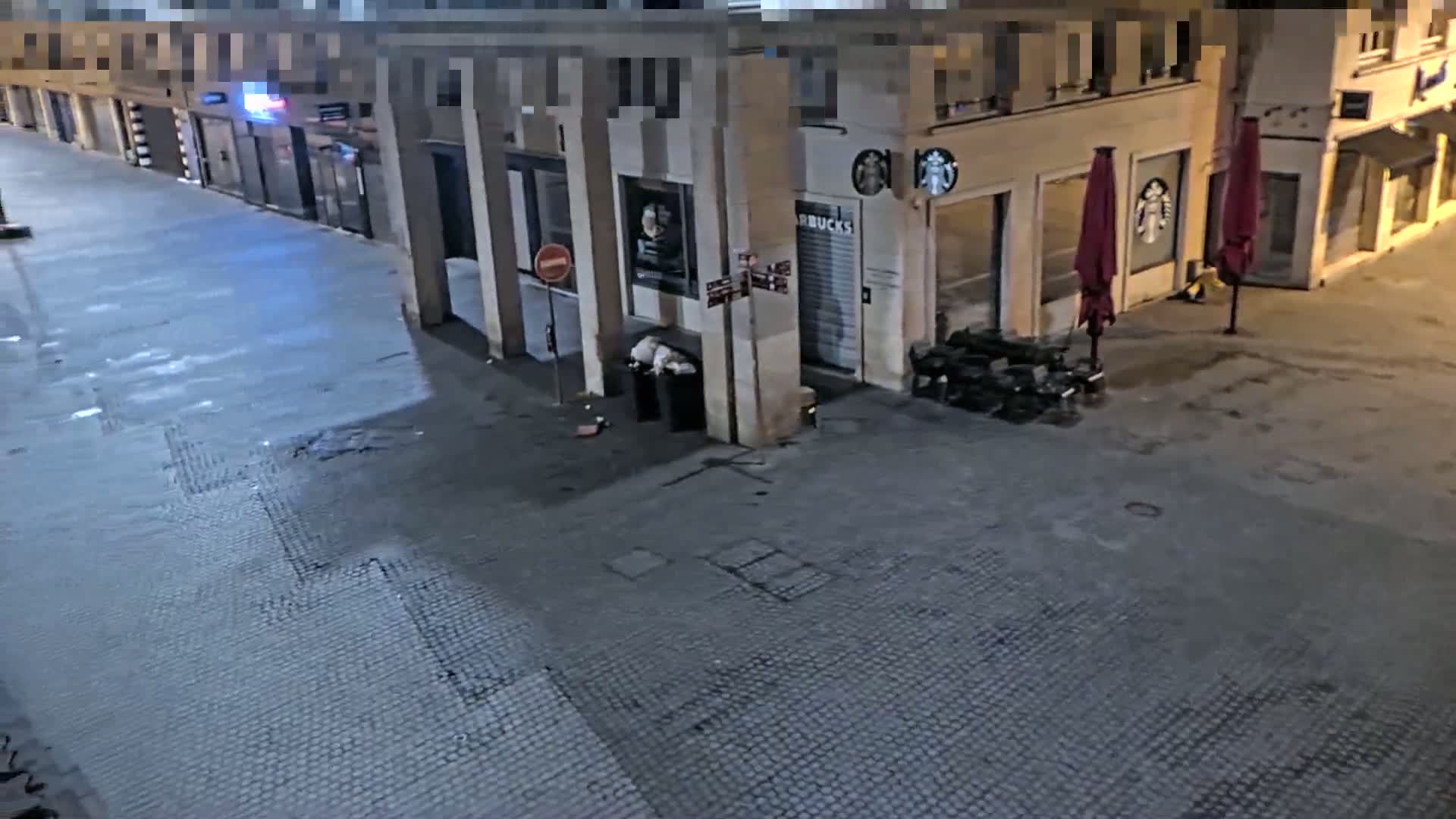 An empty street at night, showing the exterior of several buildings with closed storefronts and a few closed umbrellas outside one of them.