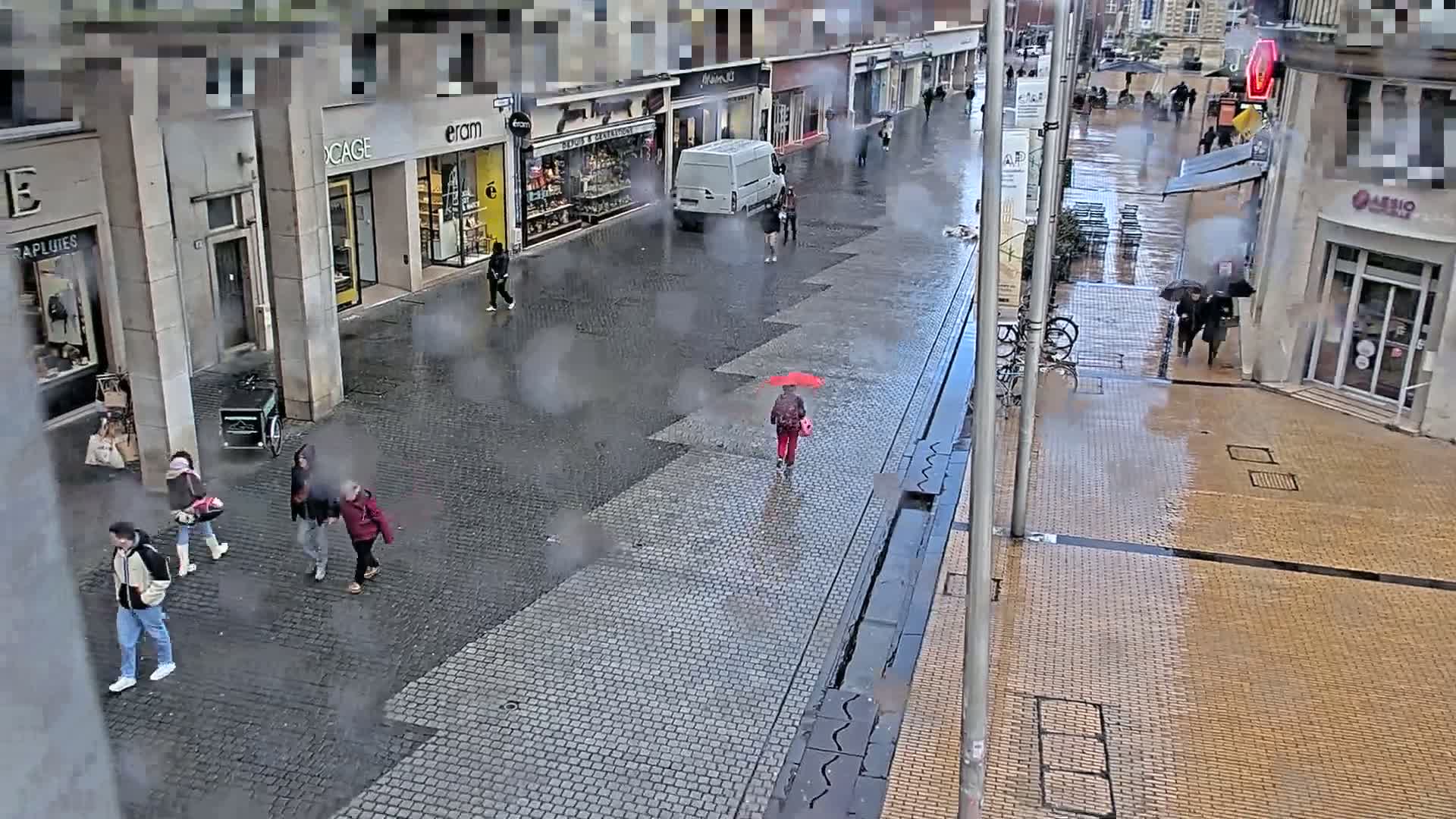 A rainy city street scene shows several pedestrians walking past shops and buildings, some carrying umbrellas.