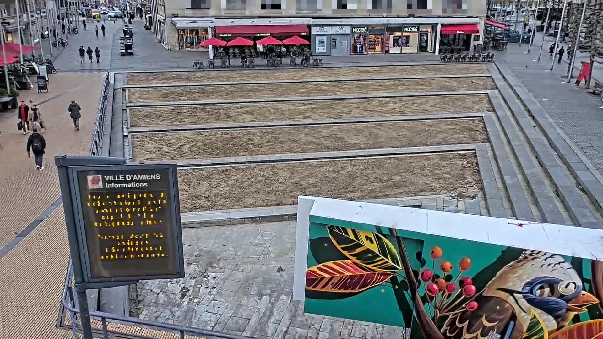 A mostly empty paved plaza with several long, rectangular, bare dirt patches, overlooked by shops and a mural of a bird and plants, on an overcast day.