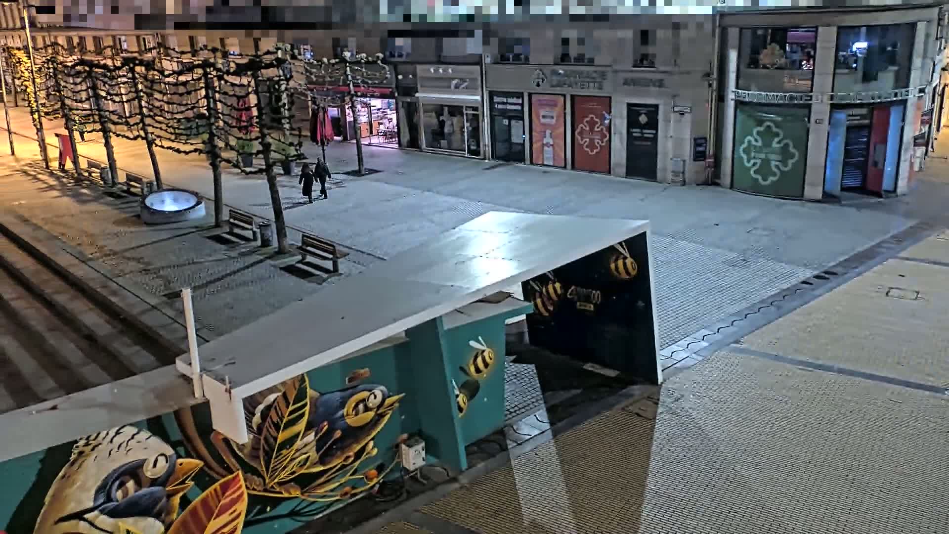 An empty, nighttime city square features a mural of birds and bees on a structure, with several bare trees lining the street and a few people walking in the distance.