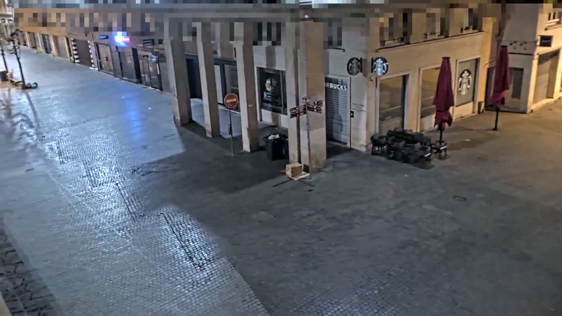 A nighttime view of a mostly empty cobblestone street with several closed storefronts, including a Starbucks, and a couple of closed red patio umbrellas.