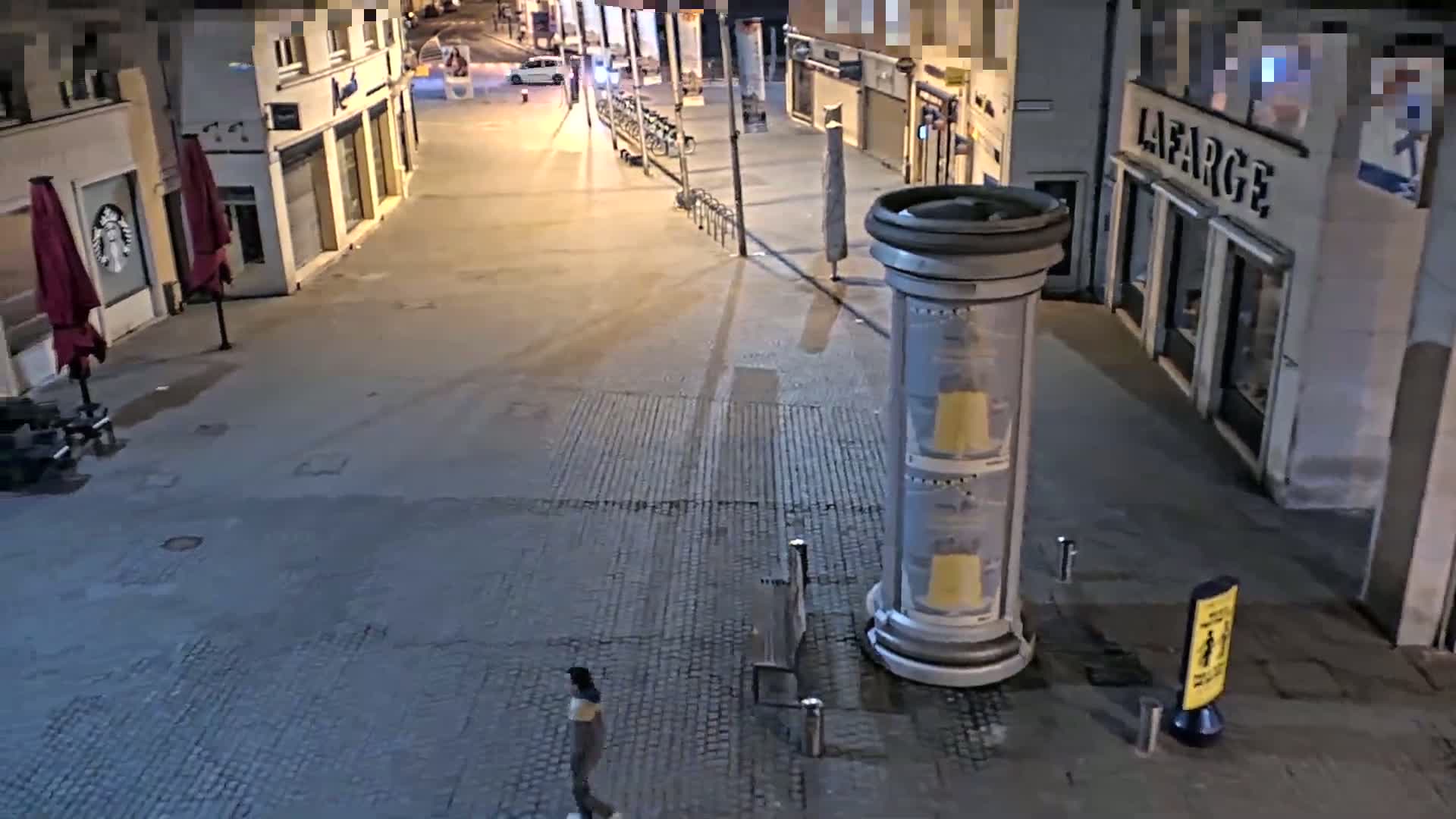 A mostly empty, paved street at night is lit by streetlights, with a single person walking and parked bicycles visible; a large cylindrical advertising column stands near the bottom of the frame.