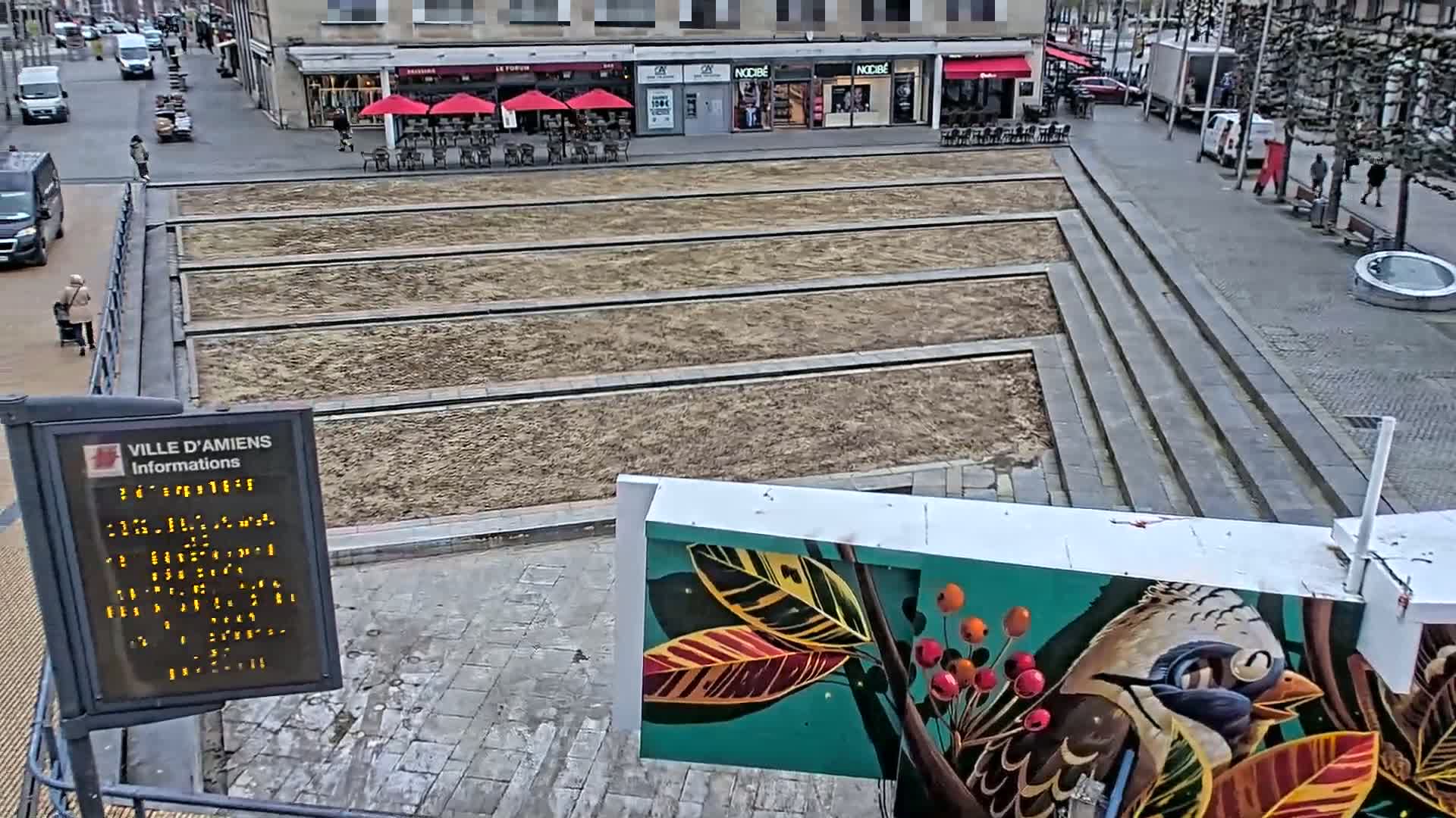 A city square with an overcast sky shows bare, rectangular planting beds, a mural of a bird and foliage, and a digital information display.