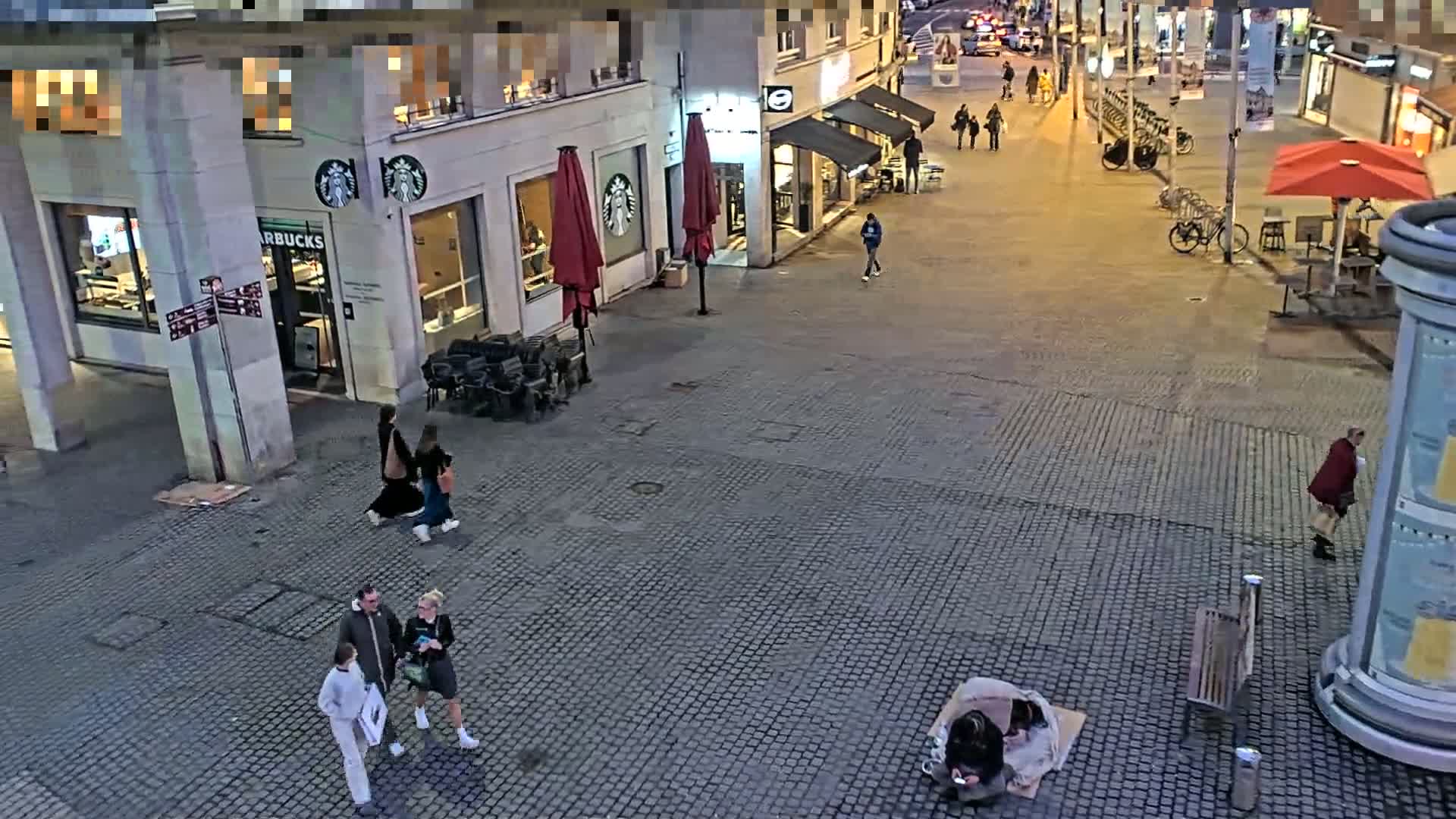 A cobblestone plaza at night, with several people walking past a Starbucks and other shops, and a person sitting on the ground.