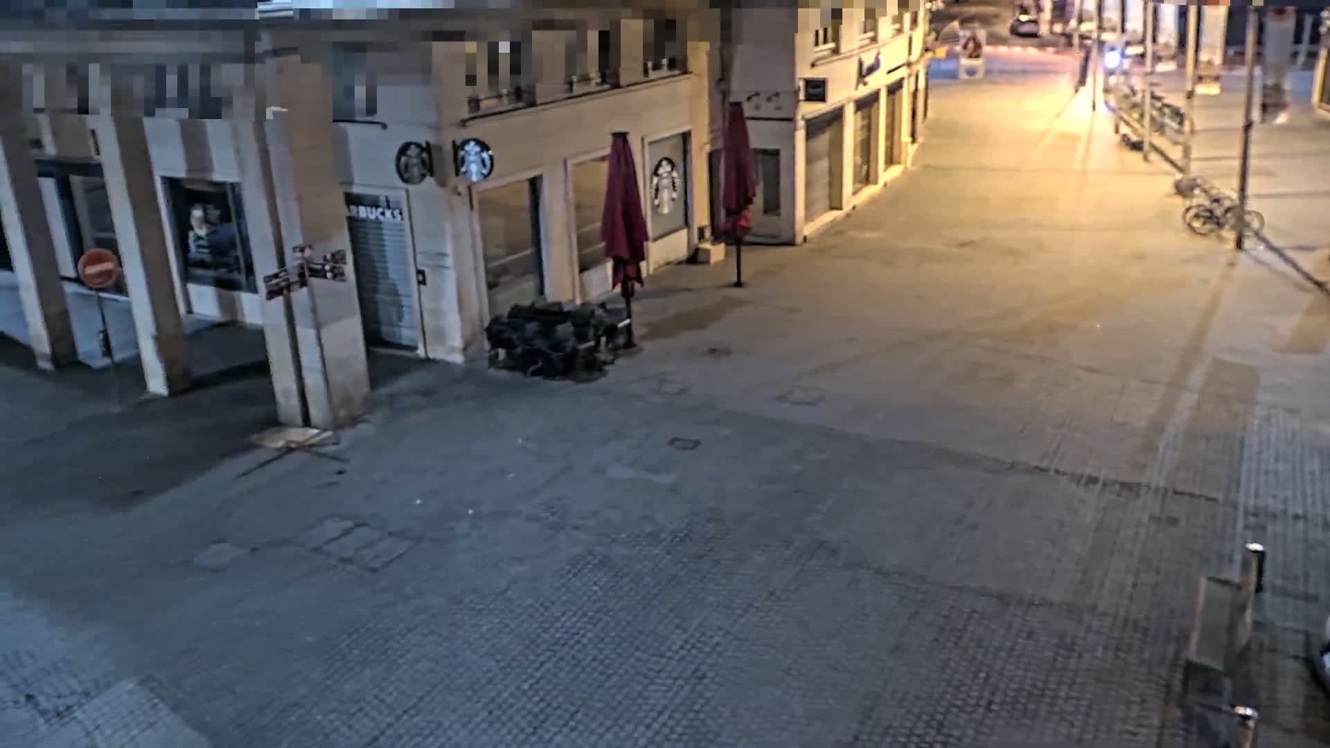 A nighttime street scene shows a mostly empty paved area in front of closed shops, with a few outdoor café umbrellas and a bicycle parked nearby.