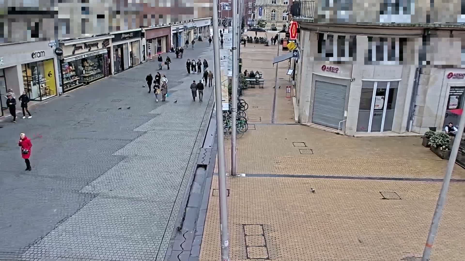 A mostly empty pedestrianized street with shops and some people walking around on a cloudy day.
