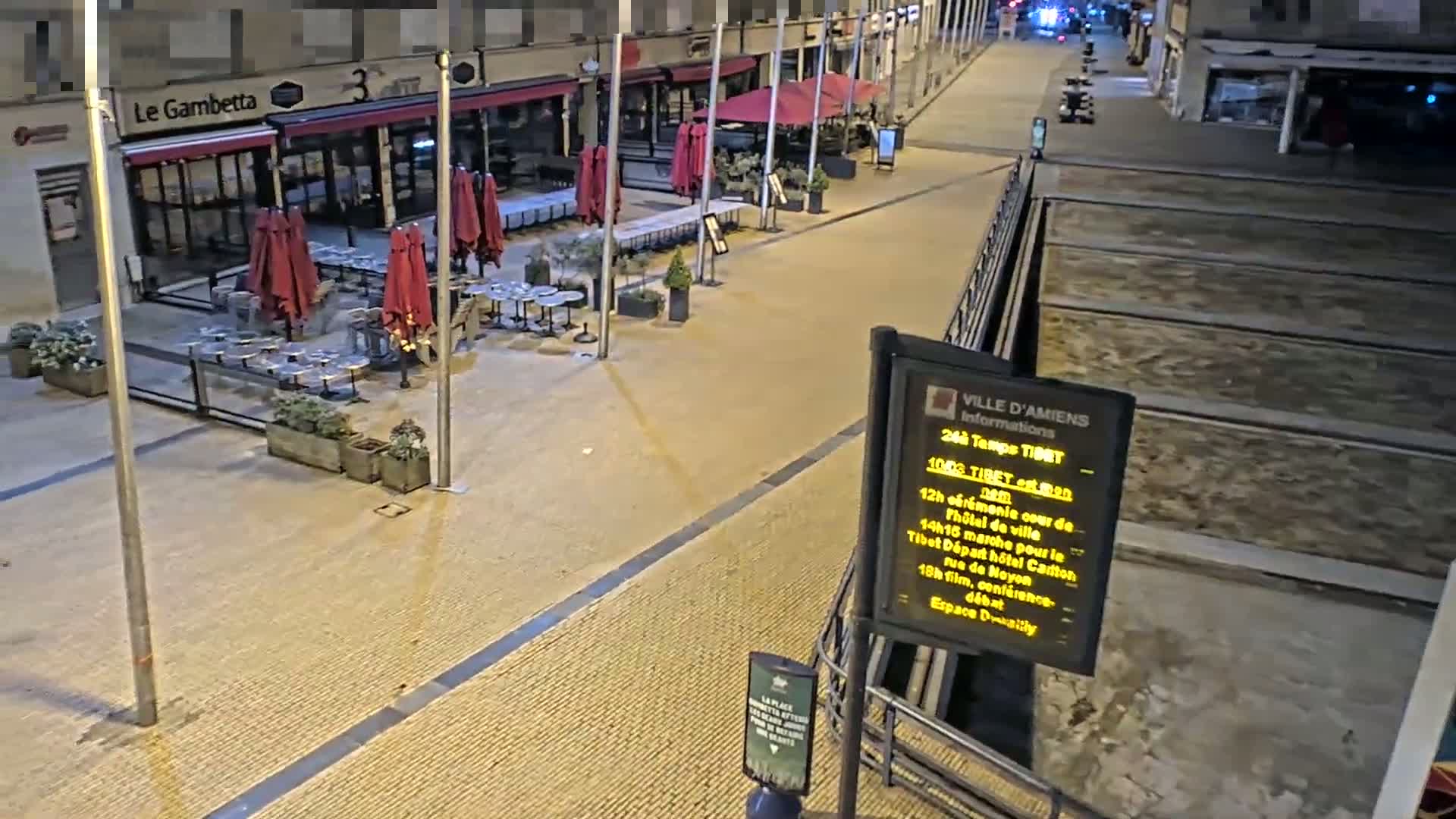 An outdoor nighttime scene shows a paved plaza with closed restaurants and their patio furniture under red umbrellas, and tram tracks running alongside.
