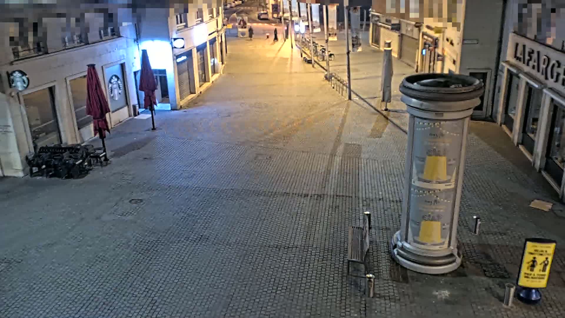 A mostly empty cobblestone street at night is lit by streetlights, with closed shops lining both sides and a few pieces of outdoor furniture scattered around.