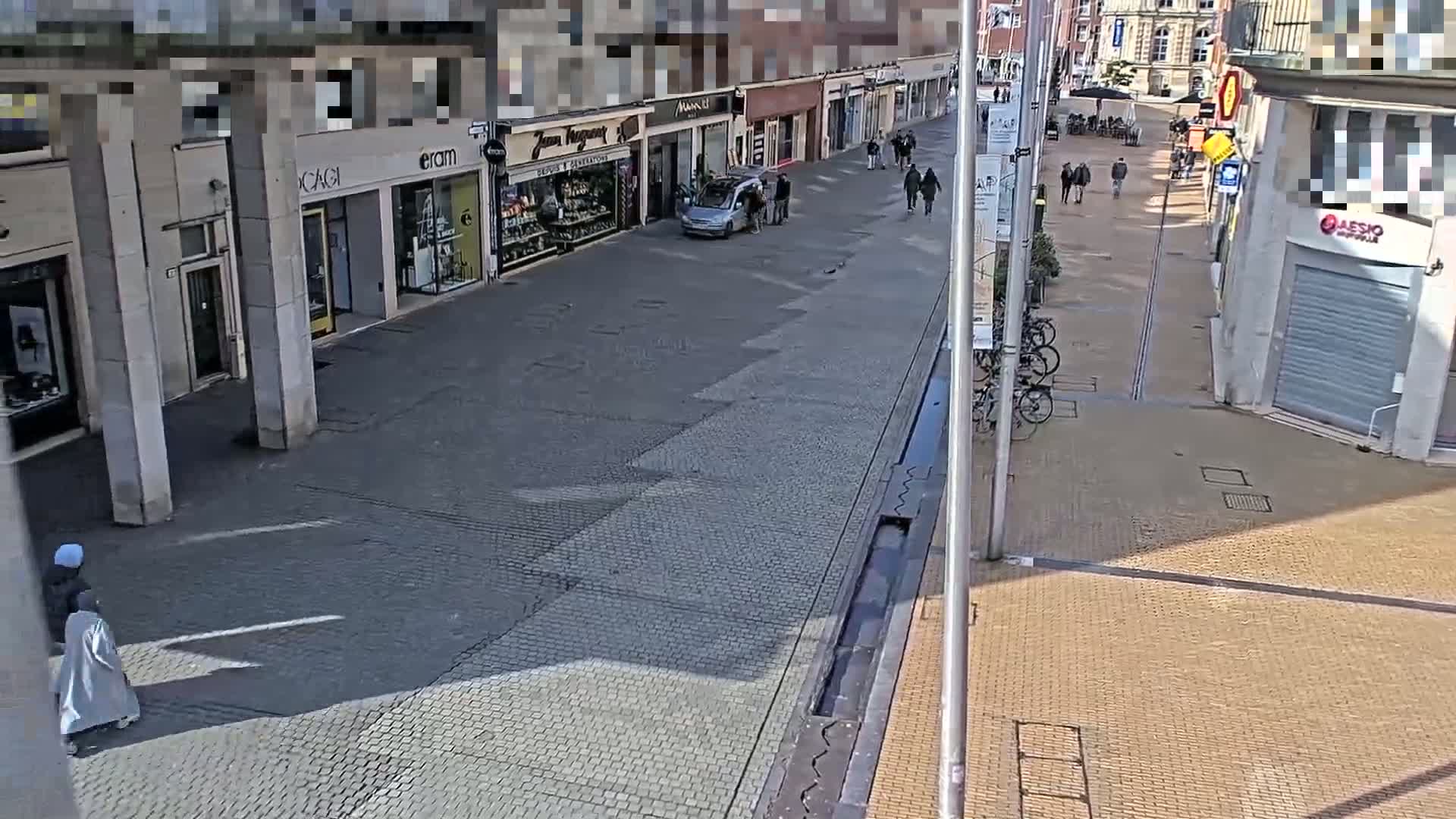 A paved pedestrian street with shops and a few people walking, under a sunny sky.