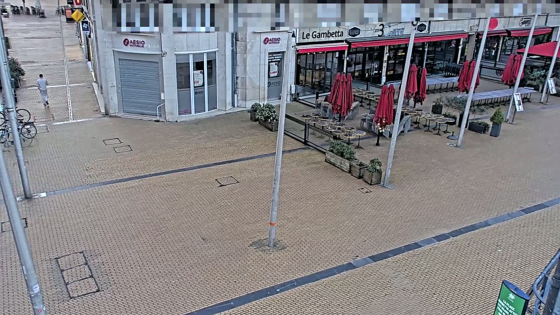 A mostly empty paved plaza with a restaurant's outdoor seating area under red umbrellas, a pedestrian walking, and several bicycles parked nearby, on an overcast day.