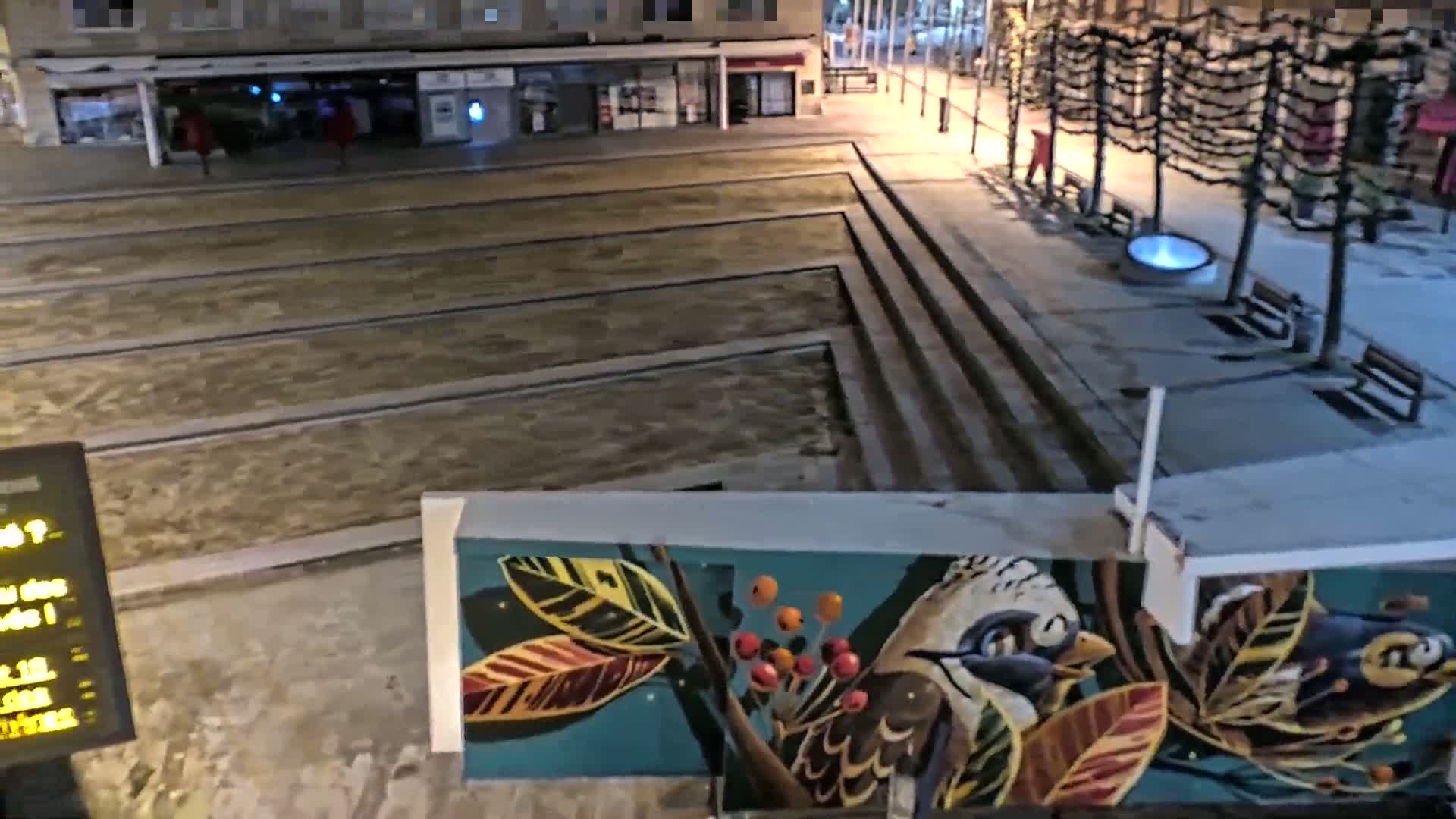 A nighttime view of a city square with recessed water features, benches, and trees, partially covered in snow, featuring a mural of birds and foliage.