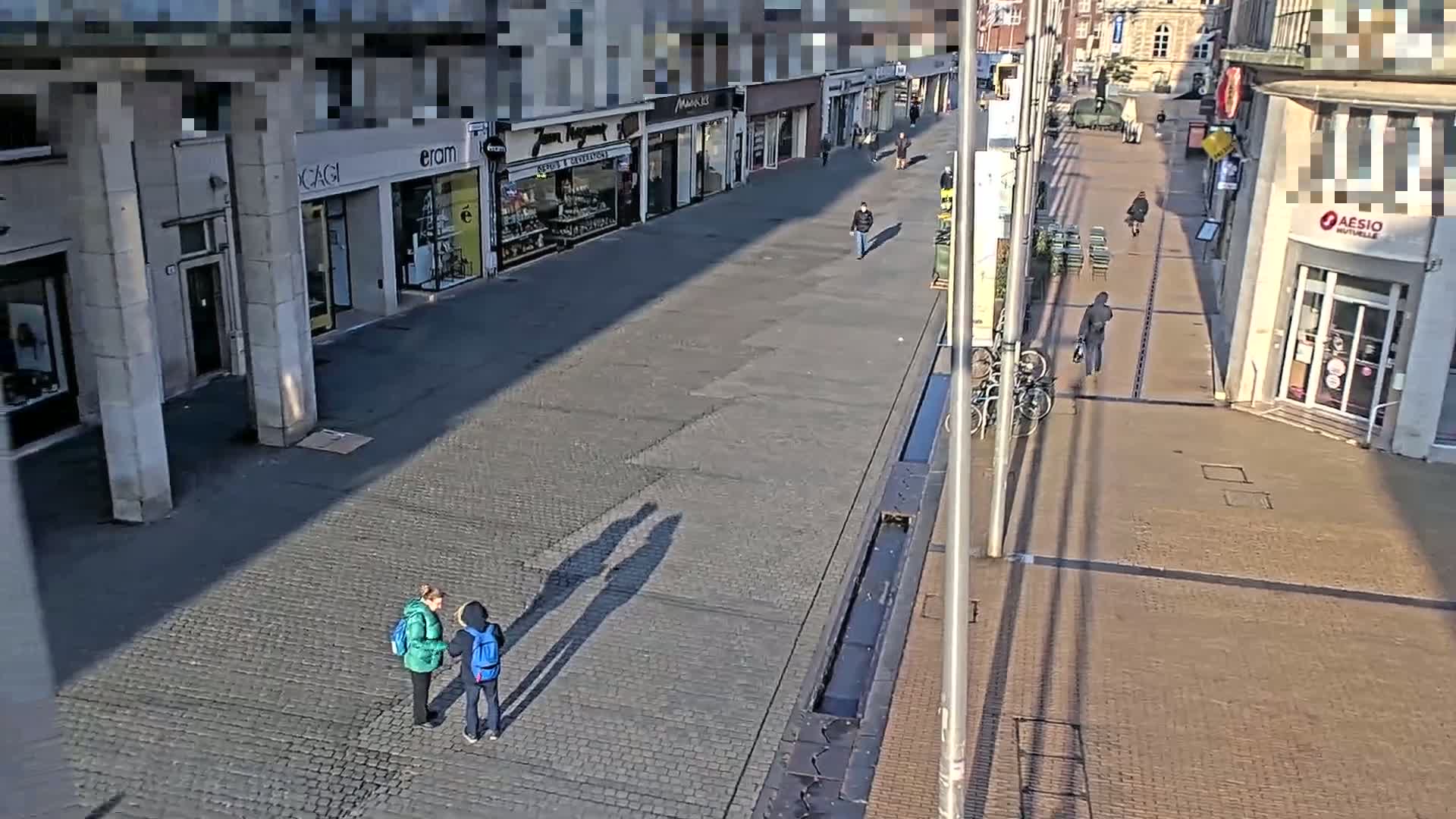 A paved pedestrian area, mostly empty except for a few people, stretches between buildings under a sunny sky; long shadows are cast from lampposts and buildings.