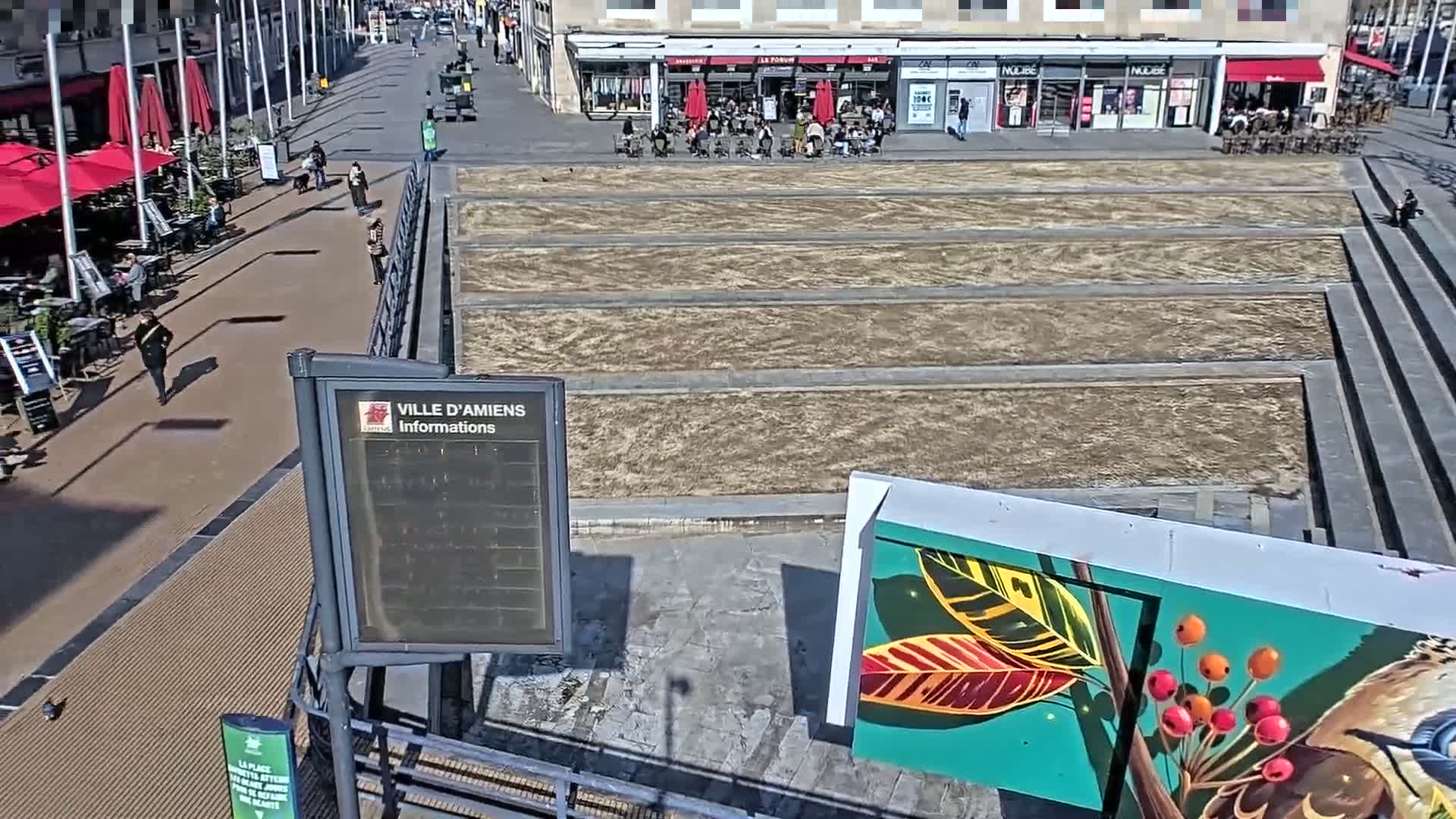 A sunny city square with a brown, barren, rectangular area,  surrounded by a paved walkway and outdoor seating areas at cafes.
