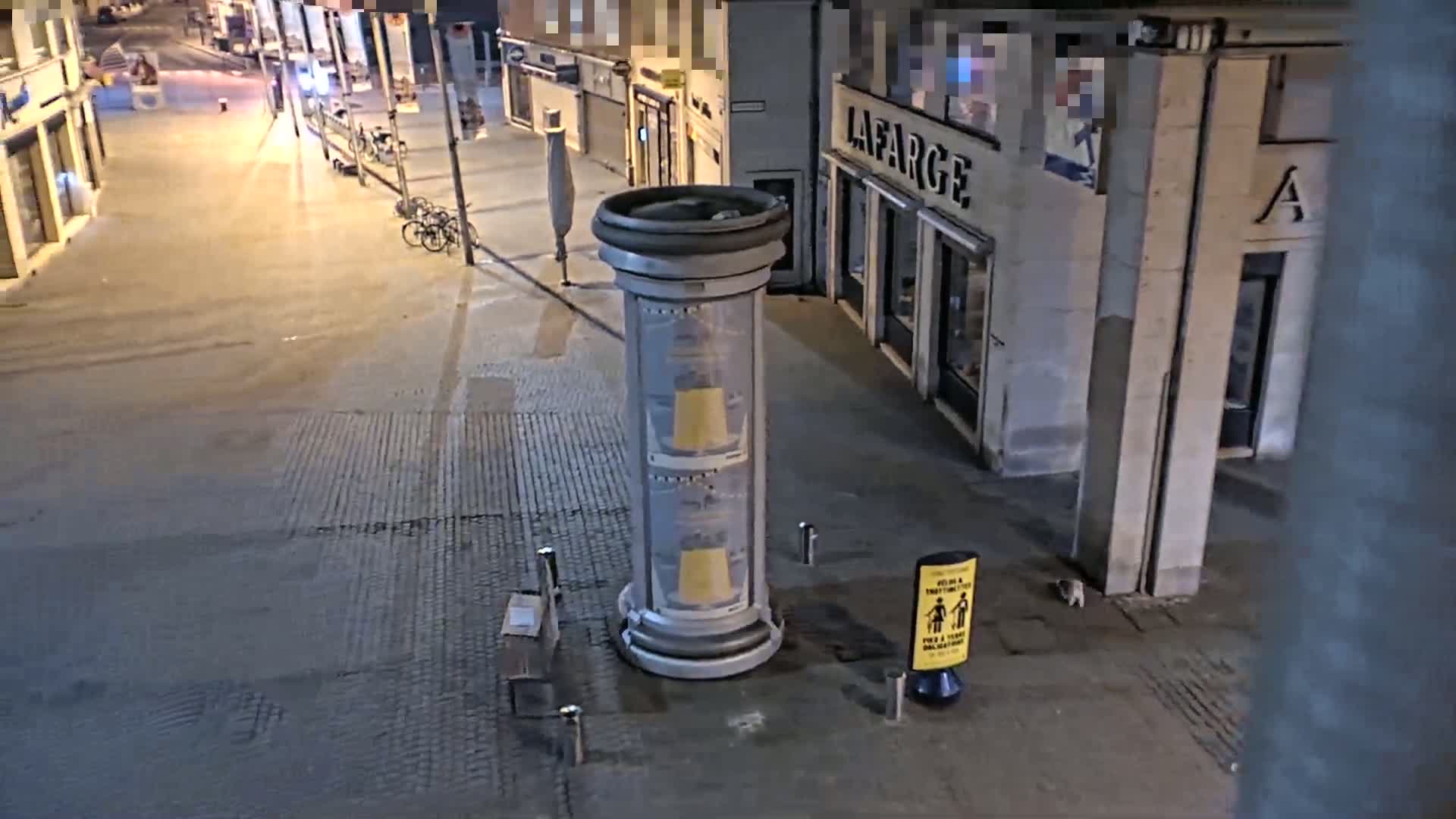 A nighttime street scene shows a cylindrical advertisement pillar on a paved area near buildings, with a few parked bicycles and a small animal visible near a social distancing sign.