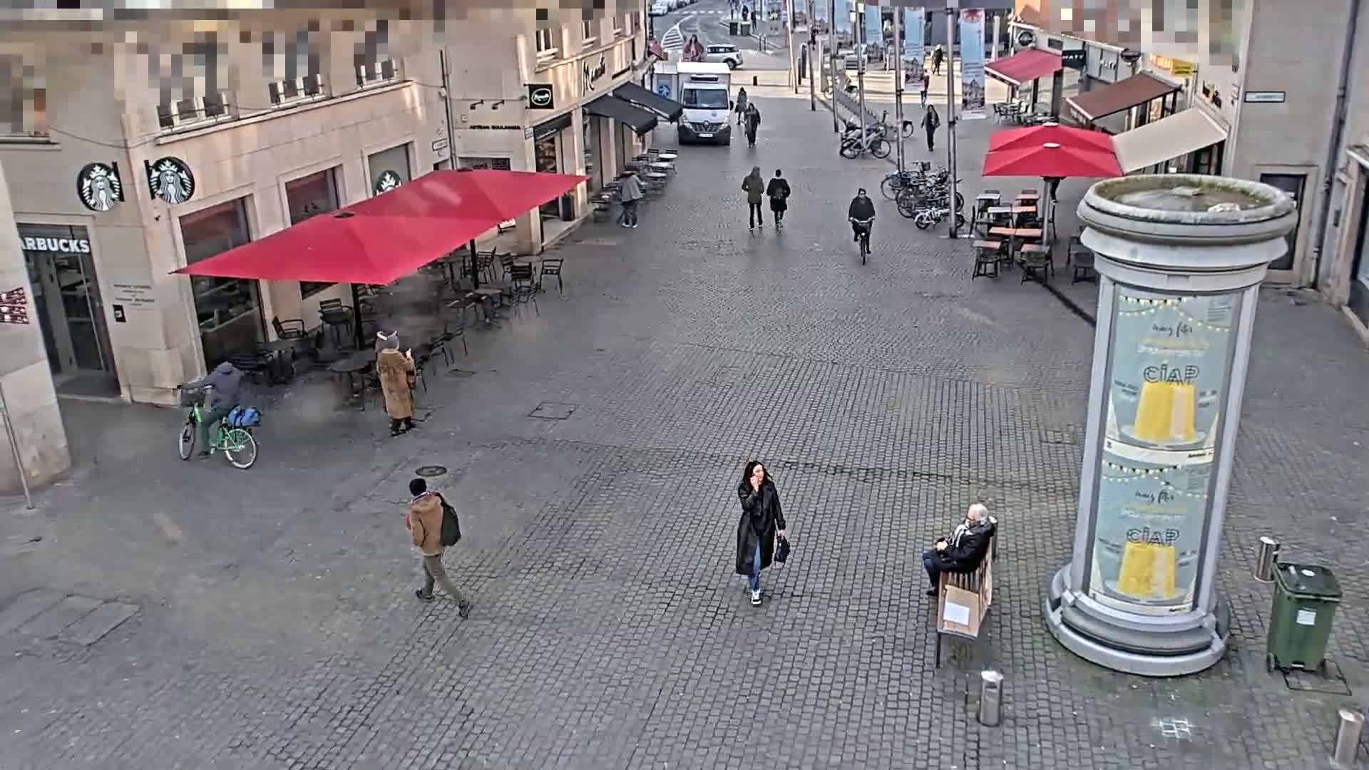 A cobblestone plaza with several people walking and sitting, outdoor seating areas with red umbrellas, and a bicycle parked near a Starbucks.