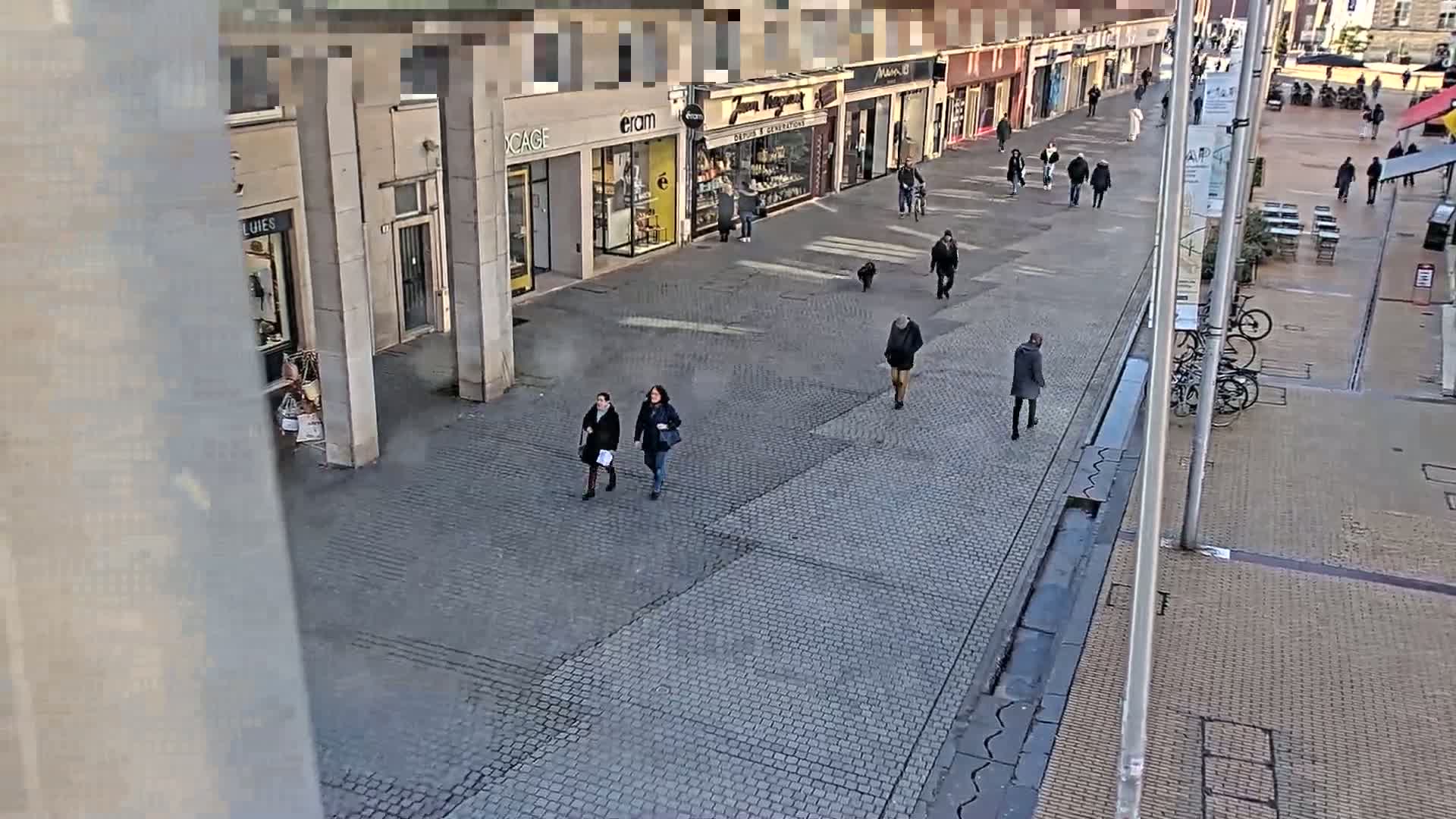 A cobblestone pedestrian walkway lined with storefronts is sparsely populated with pedestrians on a clear, sunny day.