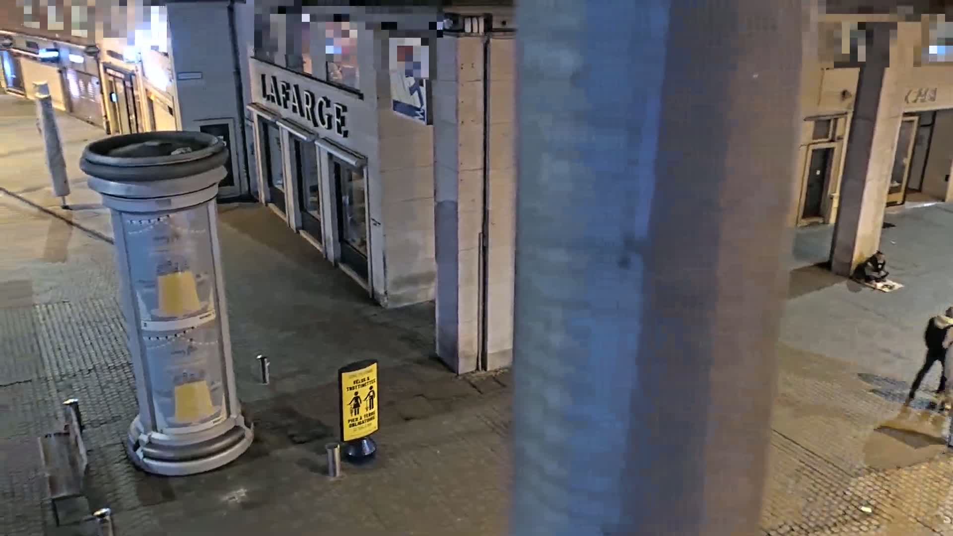 A nighttime street scene shows a person sitting on the ground near a building, another person standing nearby, and an advertising column on a sidewalk.