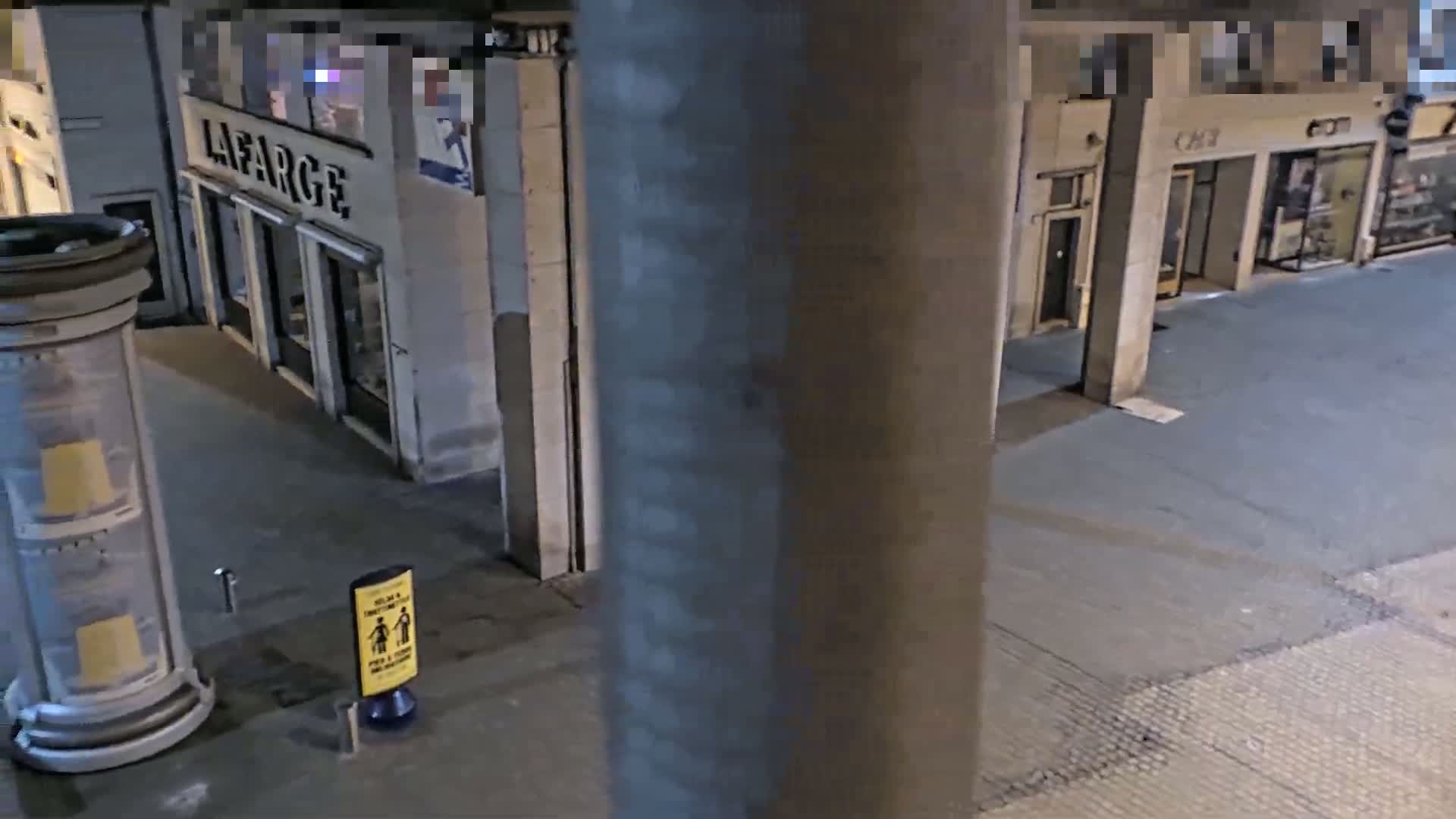 A nighttime view from a high vantage point shows a city street with storefronts, a large pillar, and a small yellow sign on the ground.