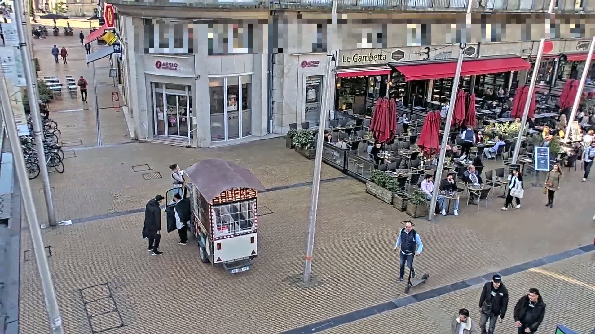 A paved city square with a food stall, several people walking and sitting at outdoor cafe tables under red umbrellas, and a few bicycles parked nearby on a partly sunny day.