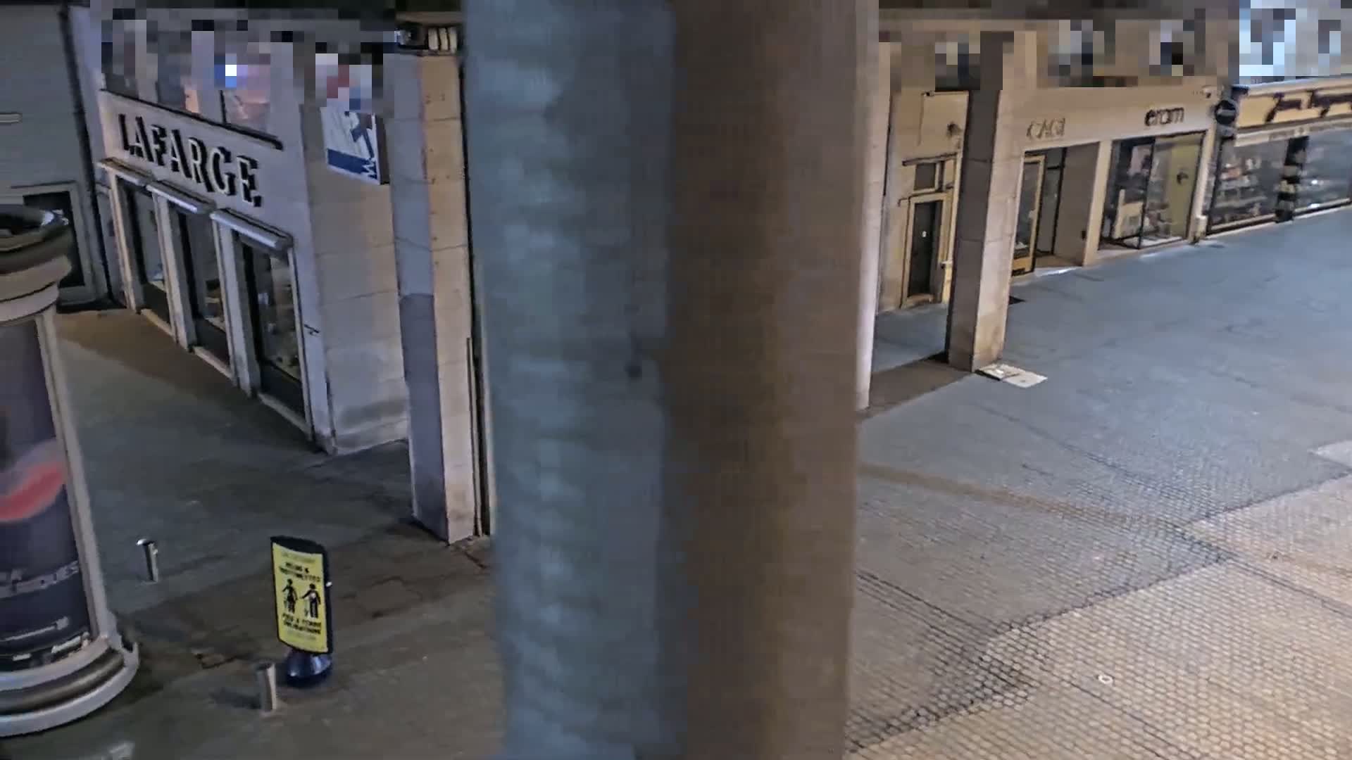A nighttime view of a city street shows a stone-paved walkway beside several storefronts.