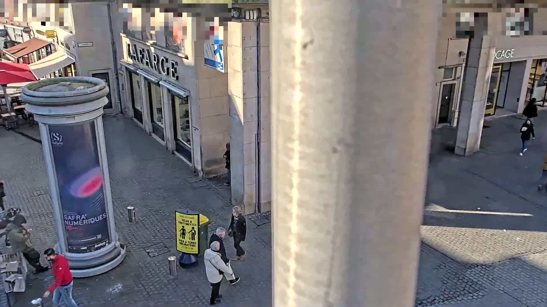A sunny city street scene shows several pedestrians walking past shops and a tall advertising column.