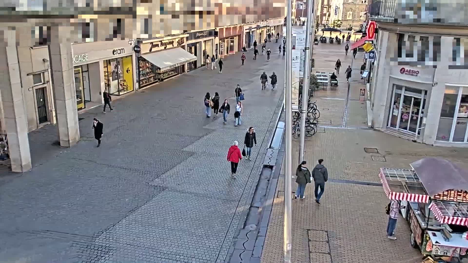 A mostly empty cobblestone street, lined with shops and pedestrians, under a partly cloudy sky.